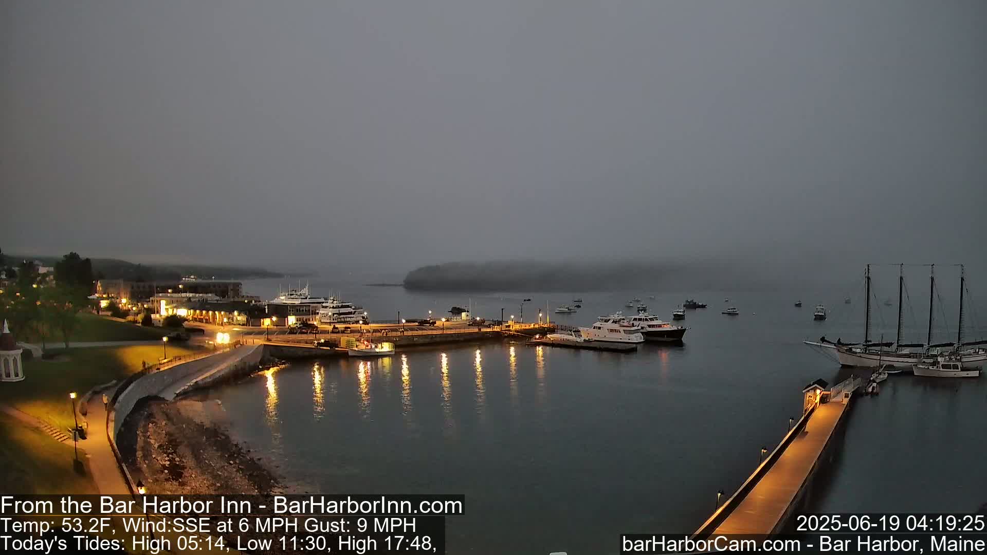 A foggy harbor at night shows a collection of boats moored at a pier, with lights illuminating the waterfront and surrounding buildings.