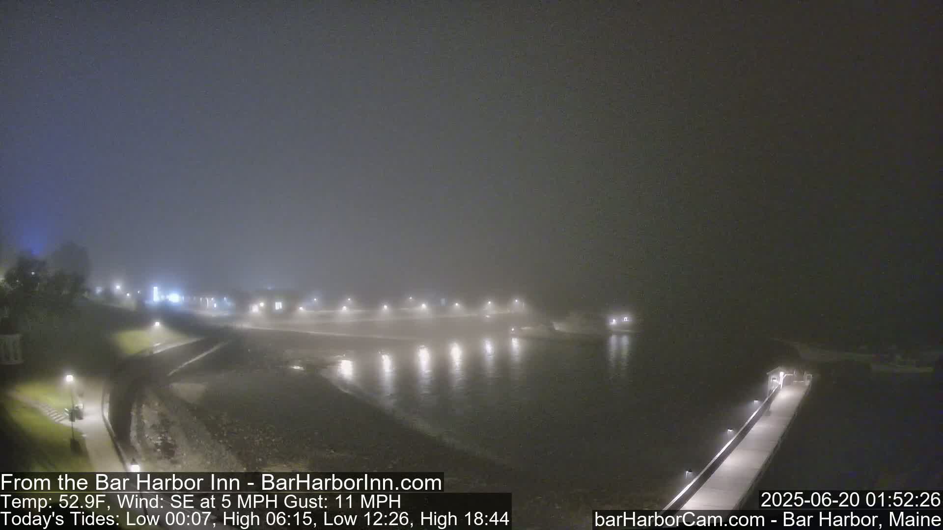 A foggy night scene shows a calm body of water with lights along a waterfront walkway and buildings in the distance.