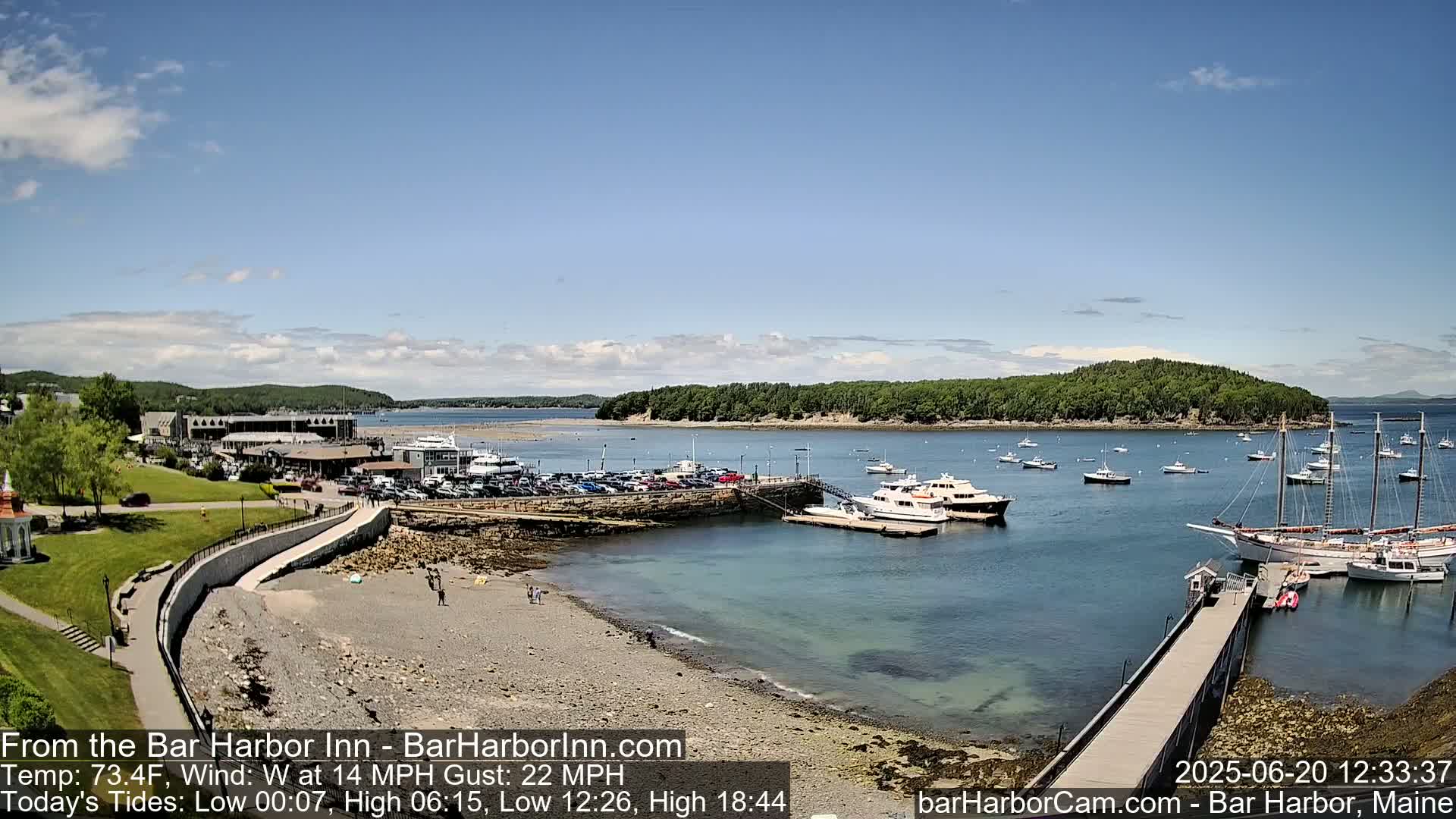 A mostly sunny harbor scene with numerous boats docked and anchored, a sandy beach, and a grassy area with buildings in the background, under a partly cloudy sky with a west wind.