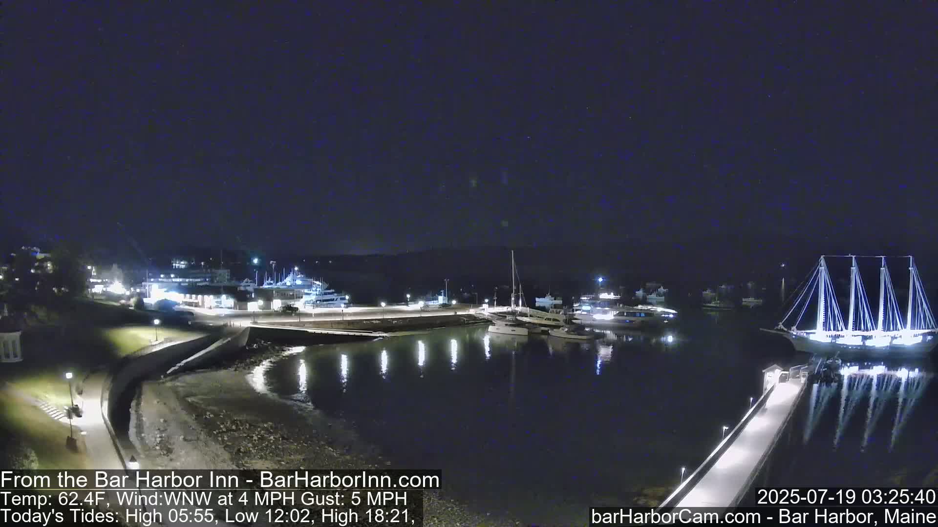 A nighttime view of a harbor with several boats and a large sailboat docked, under a clear, dark sky.