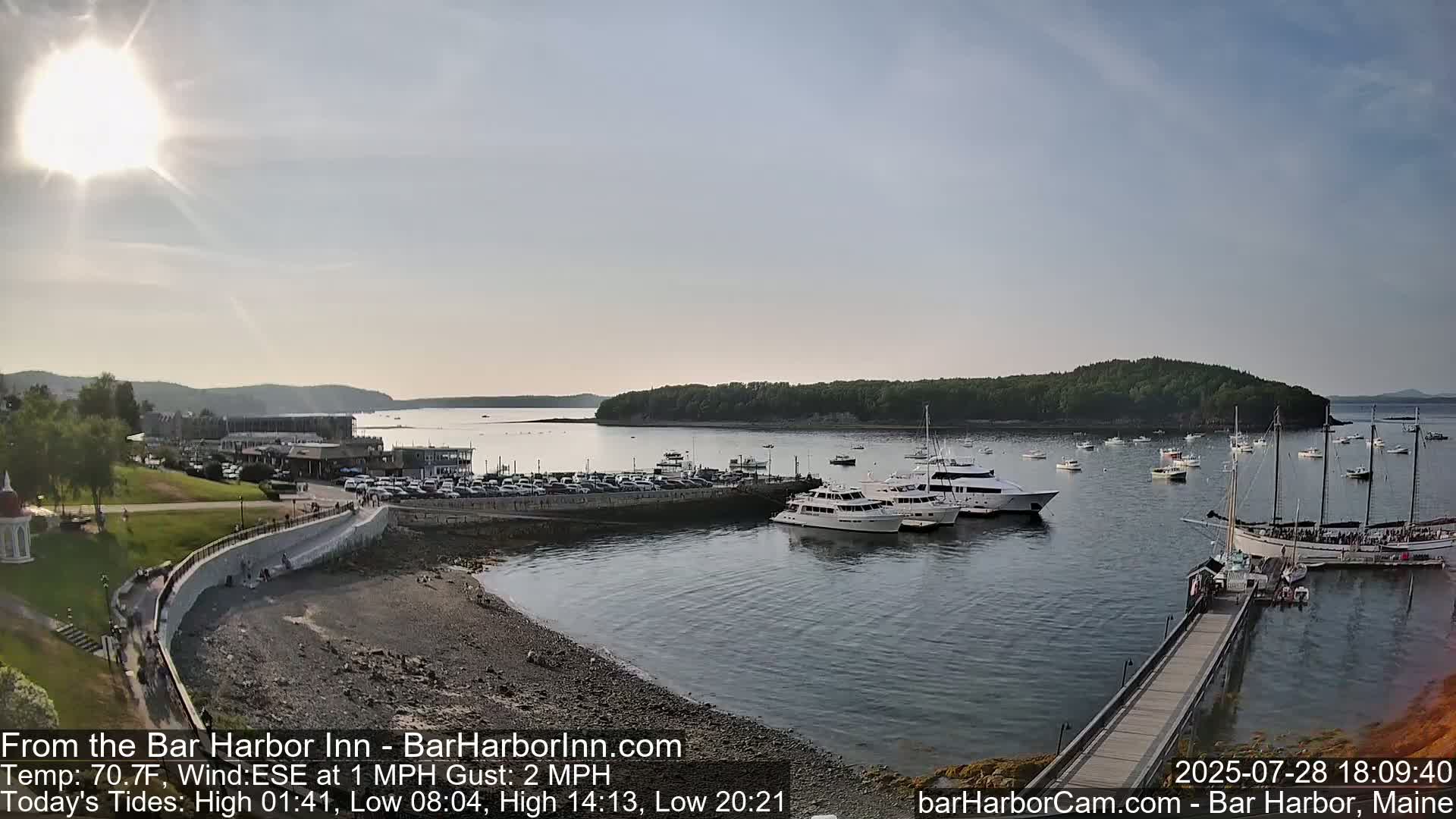 A sunny harbor scene shows numerous boats docked near a rocky shoreline with a town visible in the background.