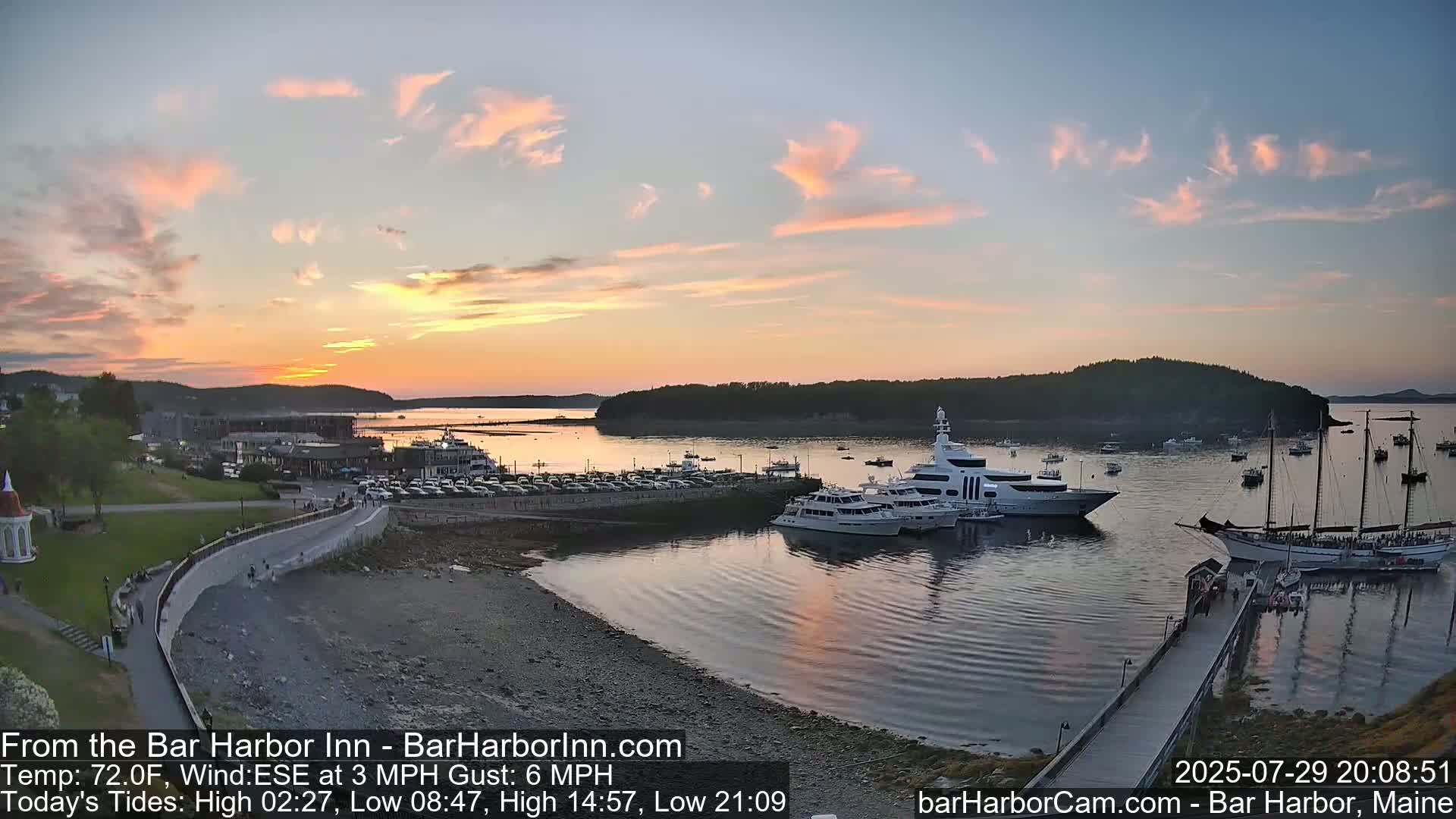 A sunny harbor scene shows numerous boats docked near a rocky shoreline with a town visible in the background.