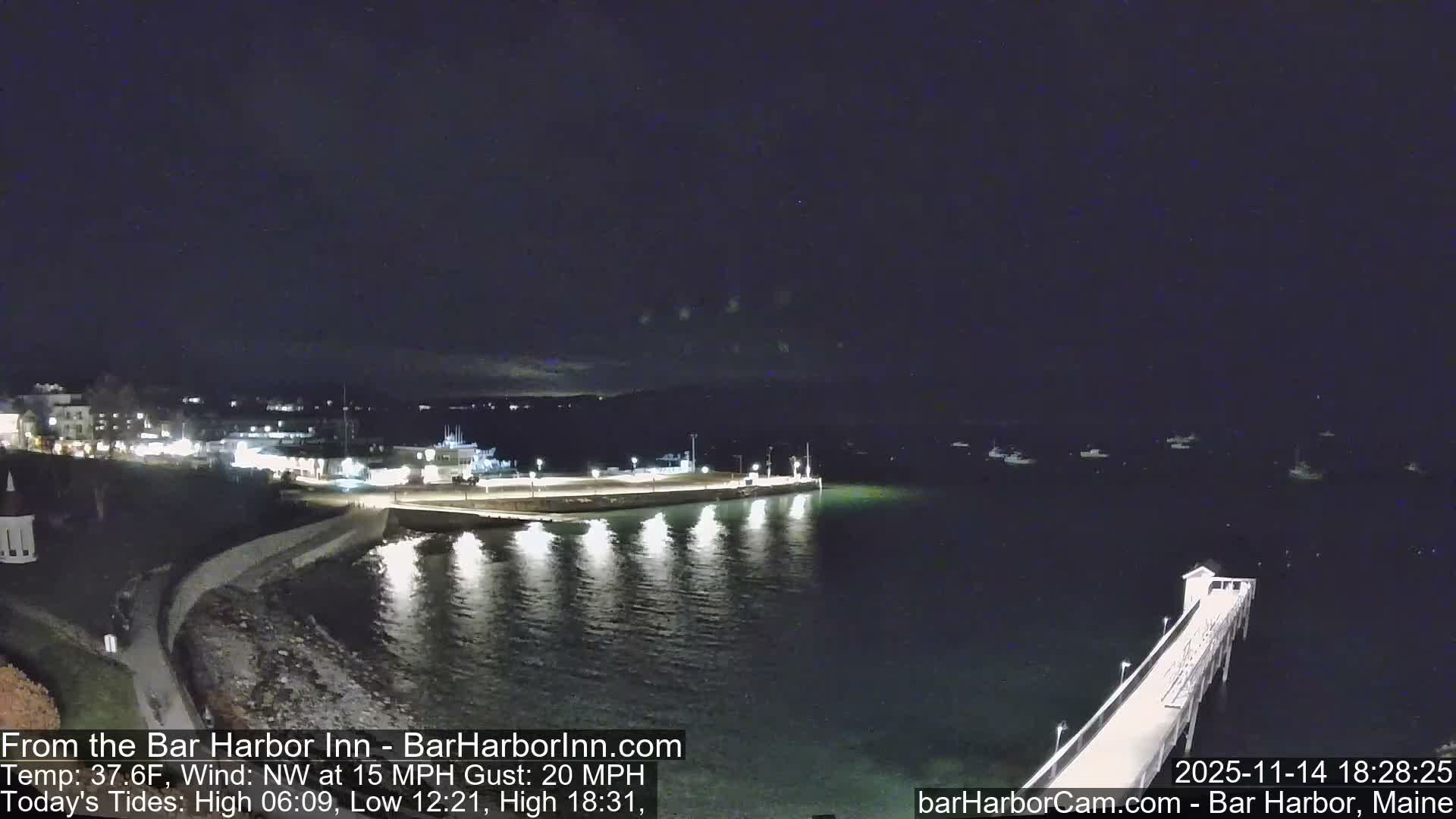 A nighttime scene captures a brightly lit coastal town and its harbor, featuring a curved causeway, a long pier with lights reflecting on the calm dark water, and several boats anchored under a mostly clear sky.