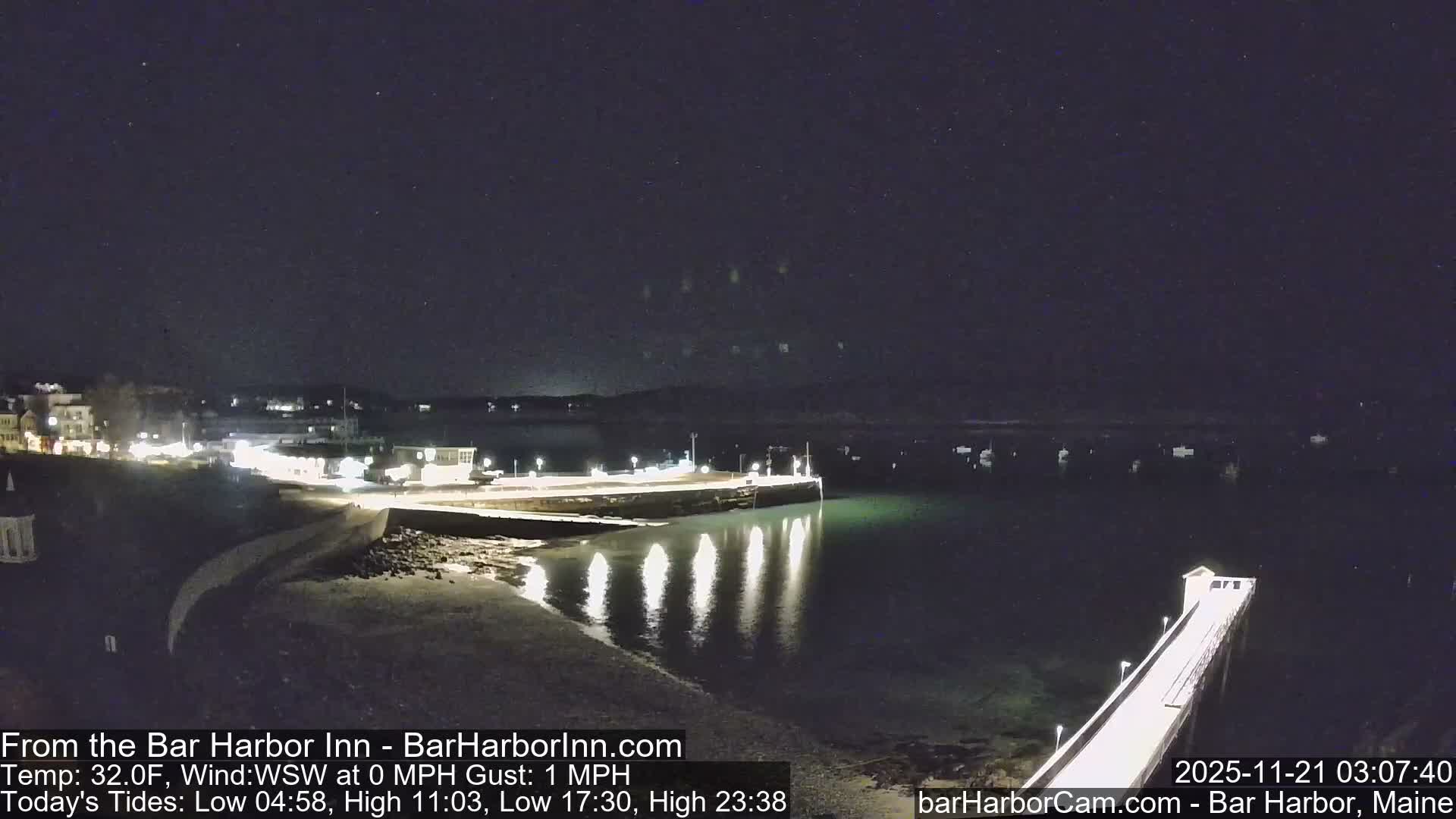 A clear, starry night reveals a brightly lit harbor with buildings and a pier reflecting on calm water, where several boats are anchored.