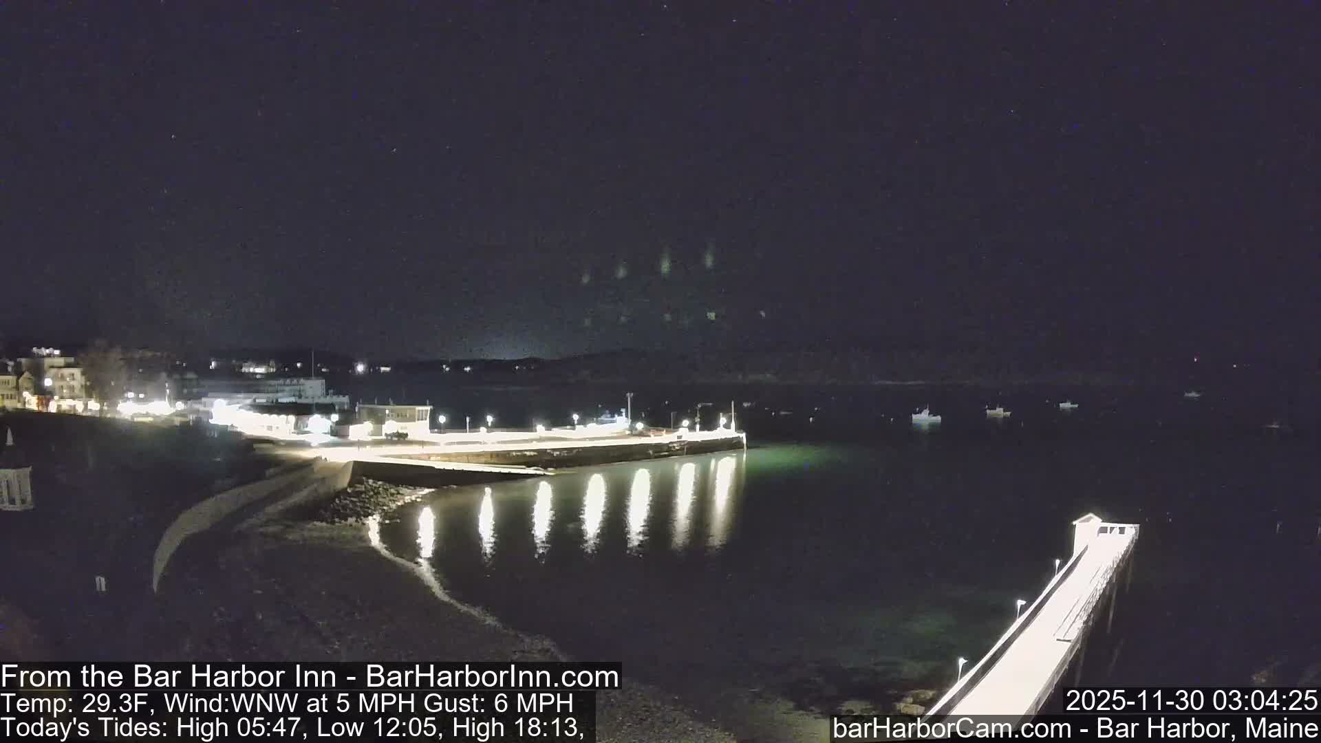 A clear and calm night reveals a brightly lit harbor with illuminated buildings and two distinct piers reflecting their lights onto the dark, calm water, which is dotted with several anchored boats.