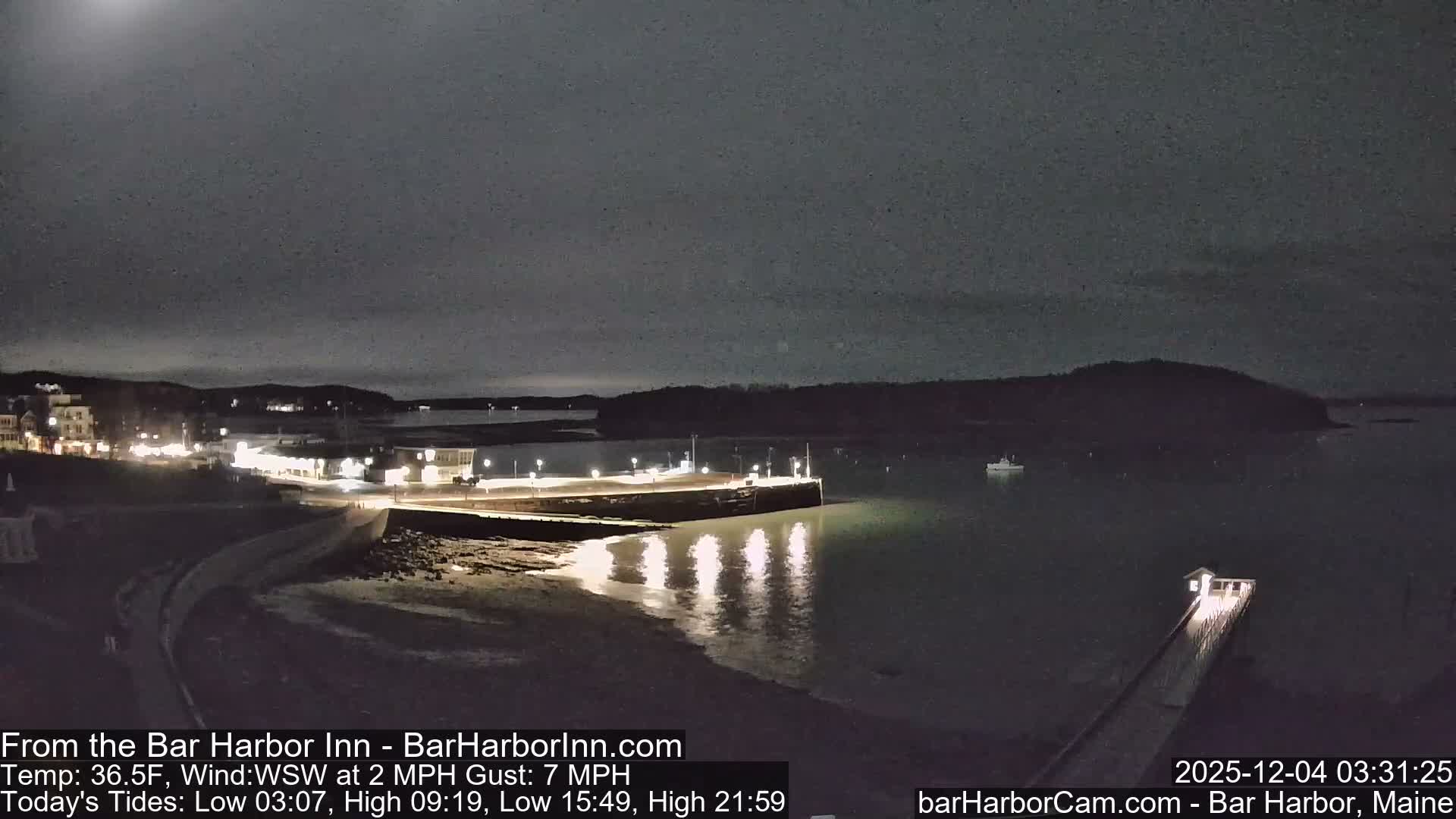 A nocturnal view of a harbor shows illuminated buildings and a well-lit pier along the waterfront, with lights reflecting on calm water and dark hills in the distance, all under a cloudy night sky.