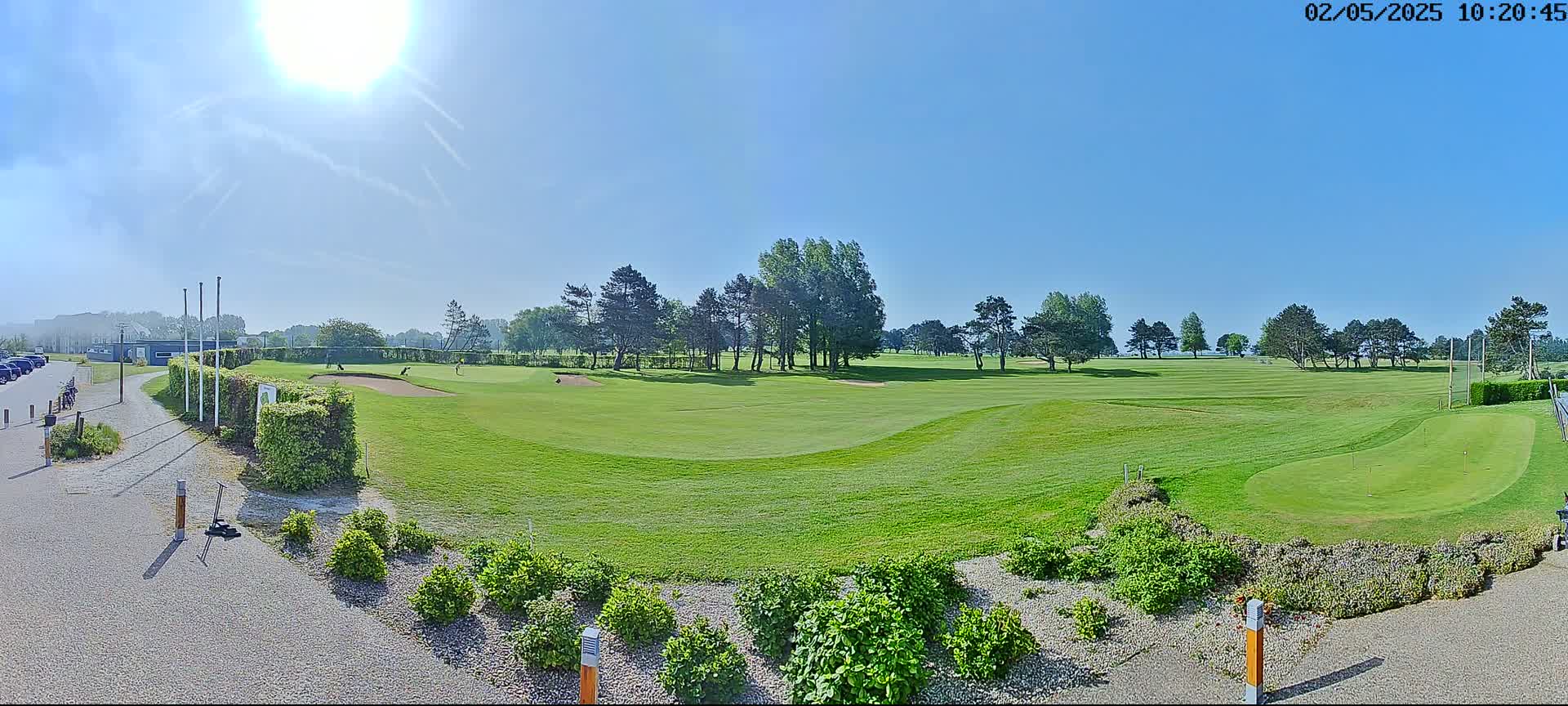 A sunny day reveals a golf course with green fairways, sand traps, and several golfers visible in the distance, bordered by neatly trimmed hedges and landscaping.
