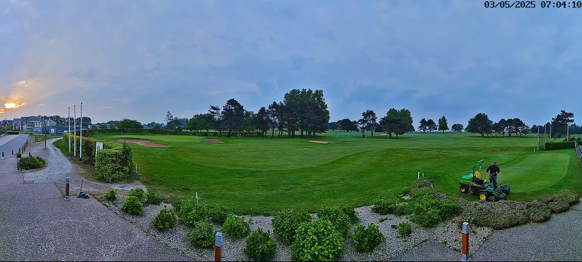 A person operates a green lawnmower on a golf course at sunrise under a mostly cloudy sky.