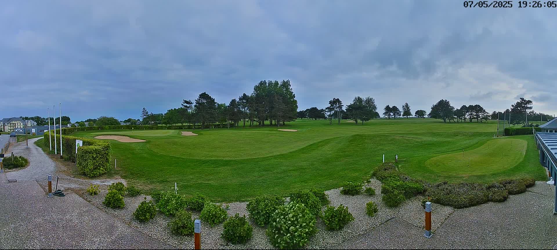 A mostly sunny golf course with a putting green in the foreground, surrounded by green landscaping and a few trees under a cloudy sky.