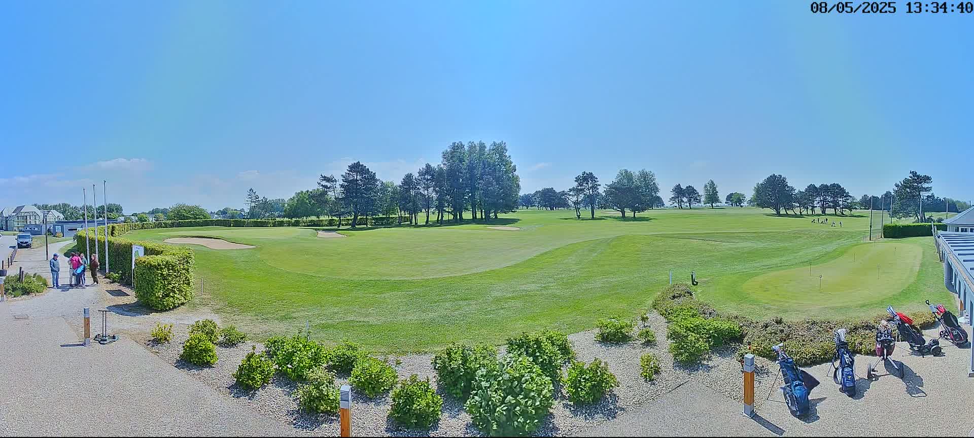 A sunny day at a golf course shows a green fairway, several golfers, and golf bags near a building.