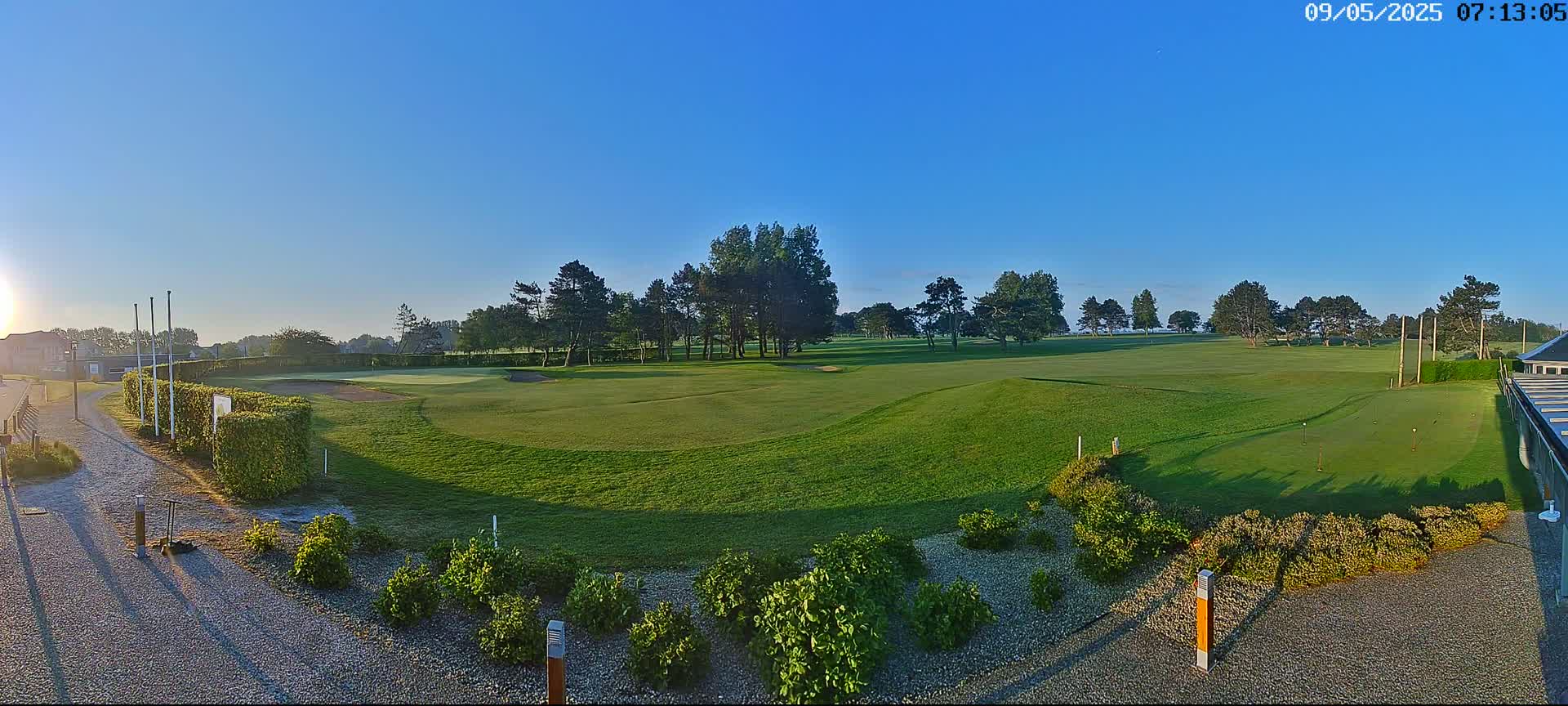 A sunny morning view of a green golf course with several trees and shrubs surrounding the putting green.