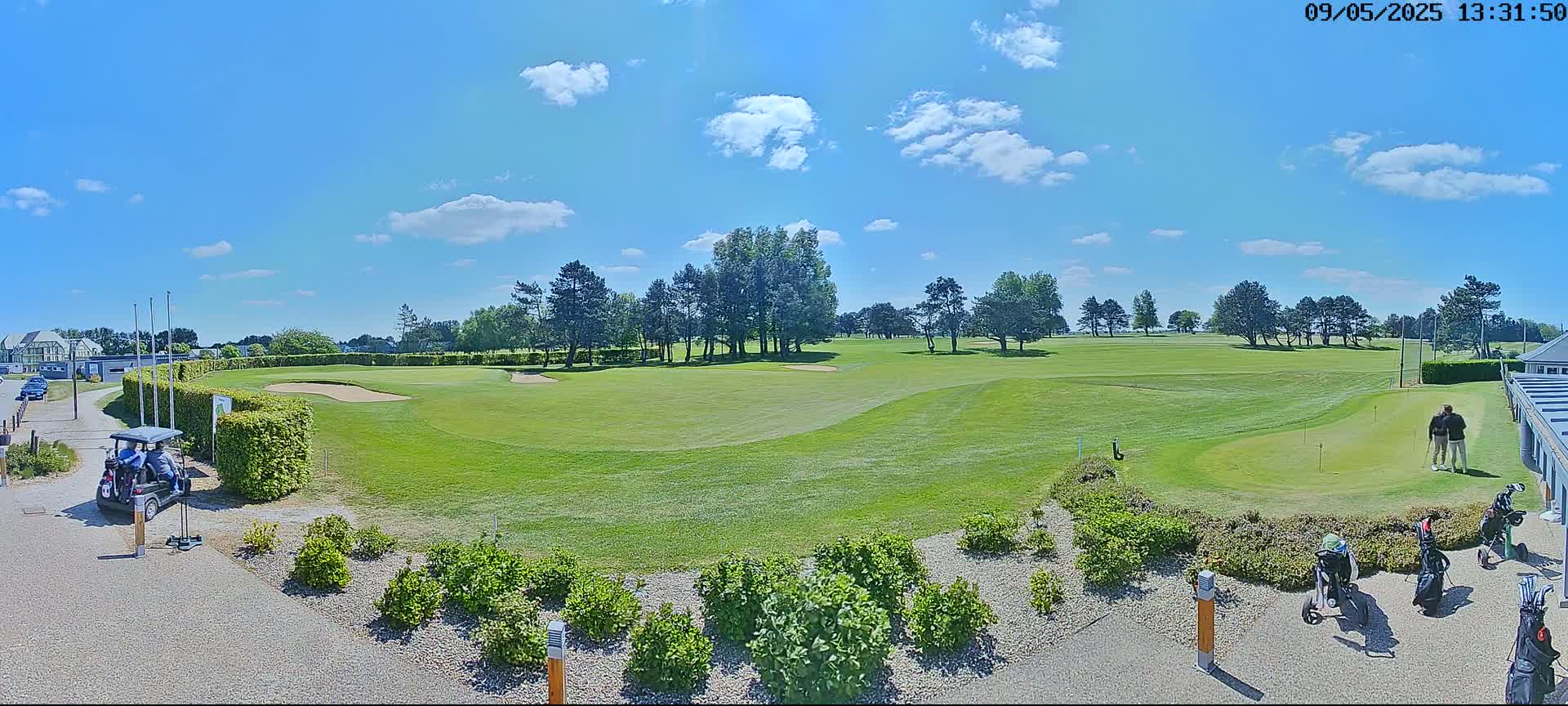 A sunny day on a golf course shows a green fairway, sand traps, several golfers, and golf carts.