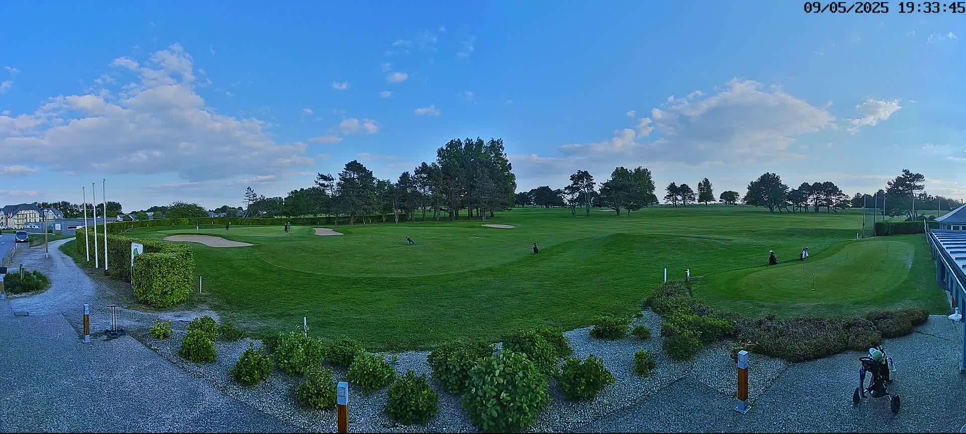 A golf course with several golfers visible under a mostly sunny sky with some clouds.