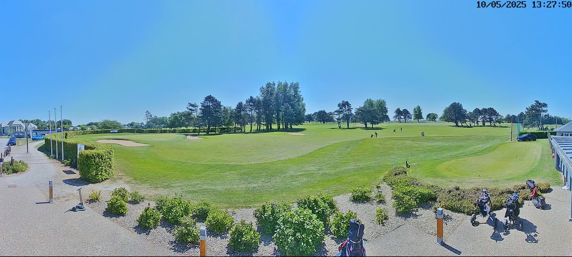 A sunny day on a golf course shows a green fairway, sand traps, and several golfers, with golf carts parked near a building.