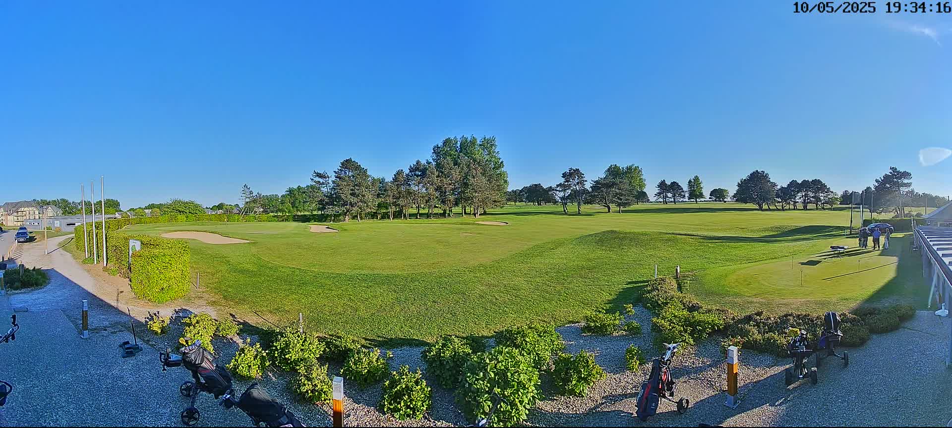 A sunny day on a golf course shows a putting green and several golf carts near a building.
