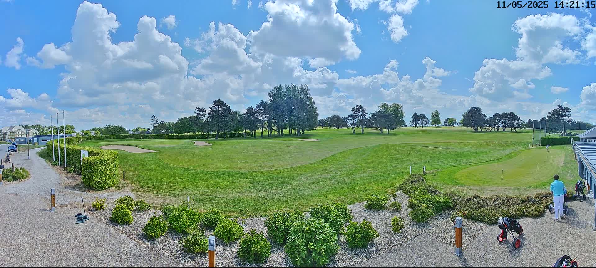 A sunny day with fluffy white clouds over a green golf course, showing a putting green and a few golfers near a clubhouse.