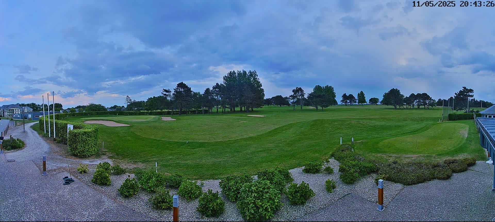 A green golf course with sand traps and a line of trees under a partly cloudy sky.