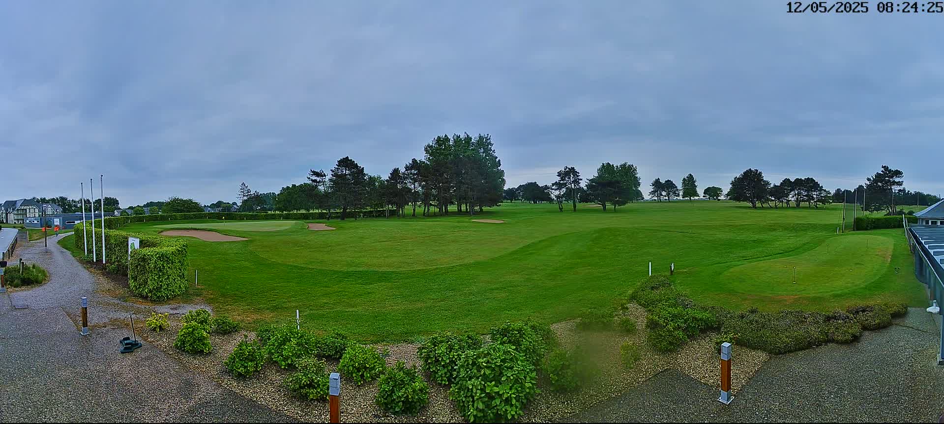 A mostly sunny golf course with green fairways and sand traps, bordered by a line of trees and shrubs, under a partly cloudy sky.