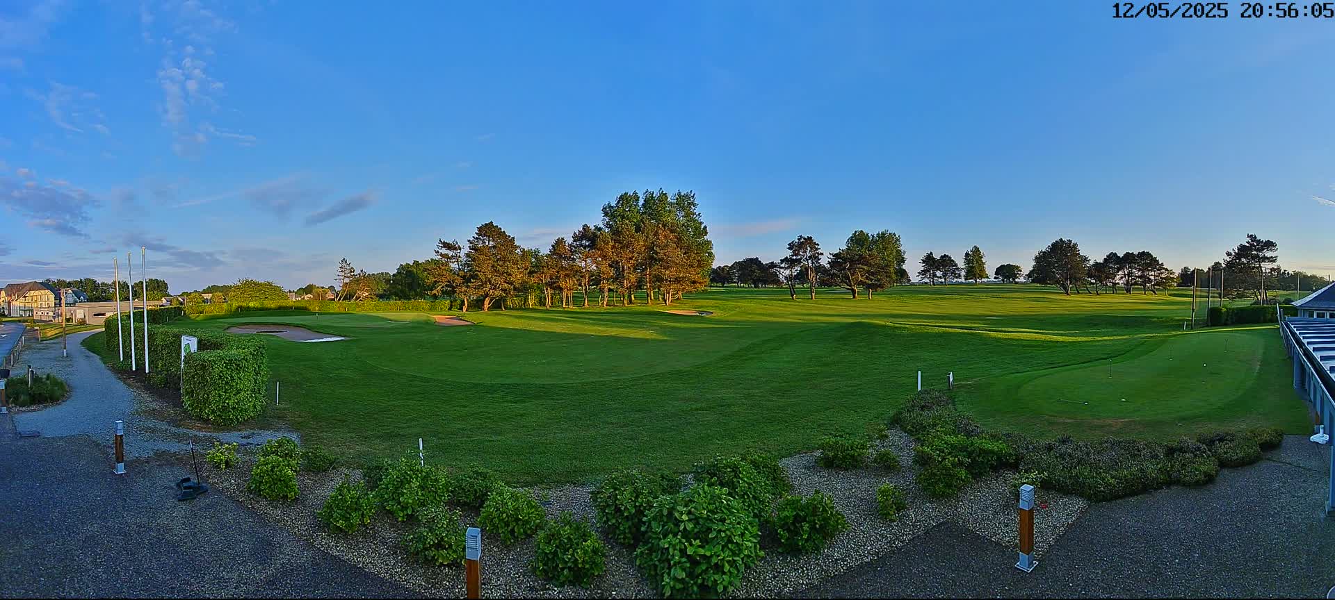 A panoramic view of a golf course at sunset under a clear blue sky shows a putting green in the foreground, with fairways and trees extending into the background.
