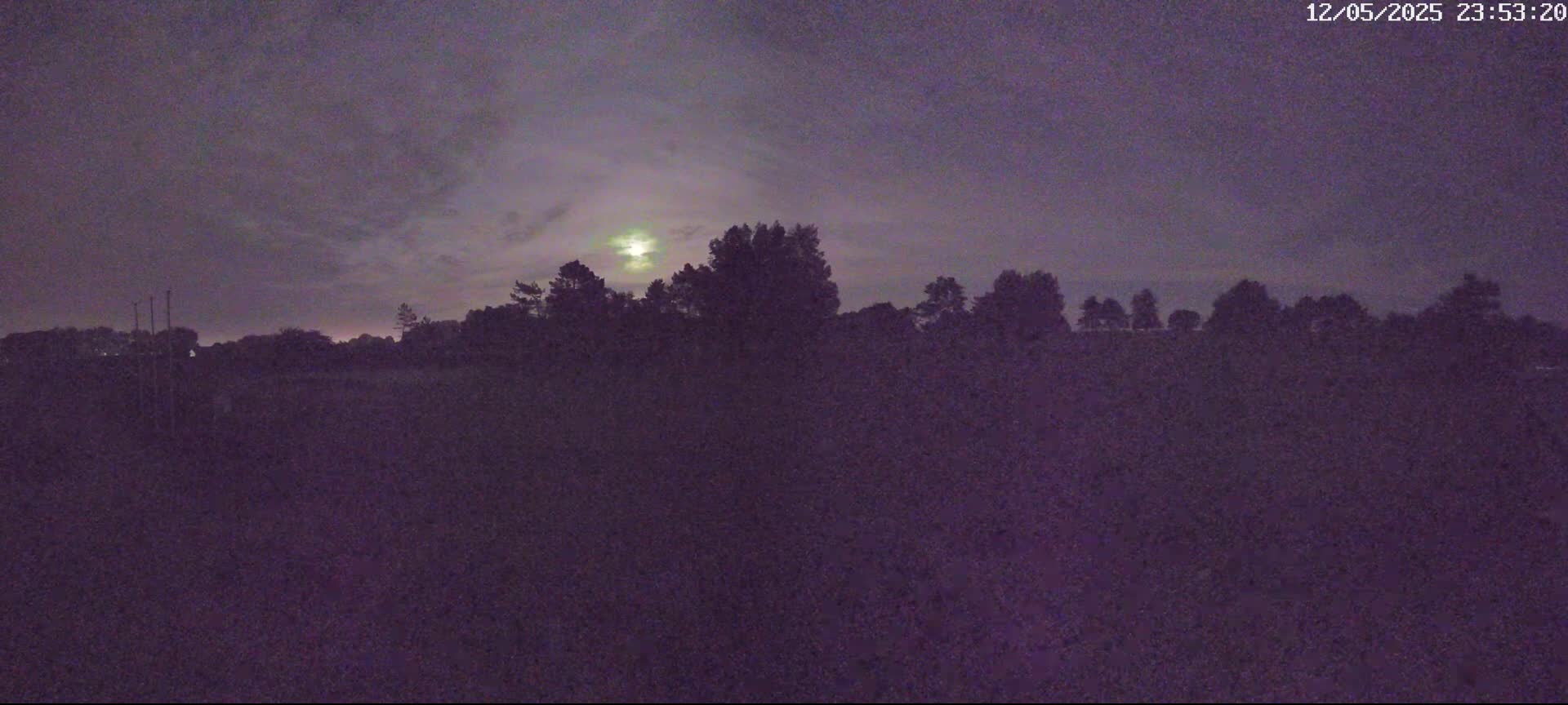 A bright moon shines through a cloudy night sky above a dark silhouette of trees and a field.