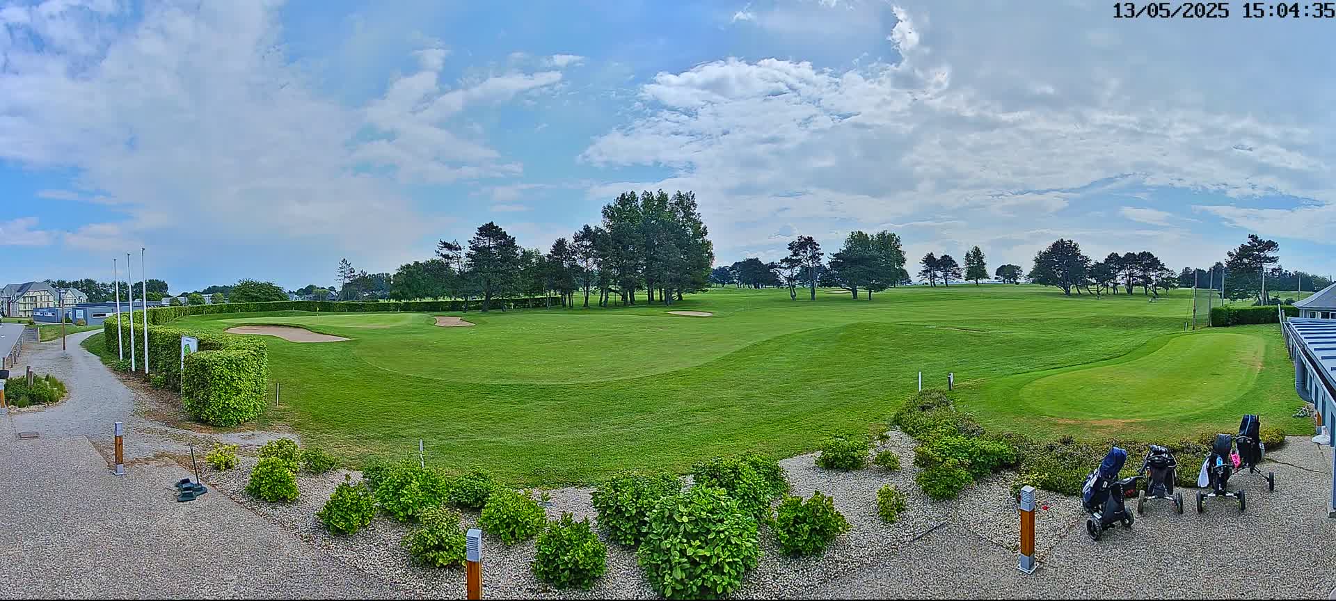 A partly sunny day reveals a green golf course with several golf carts parked near a building, bordered by lush shrubs and trees.