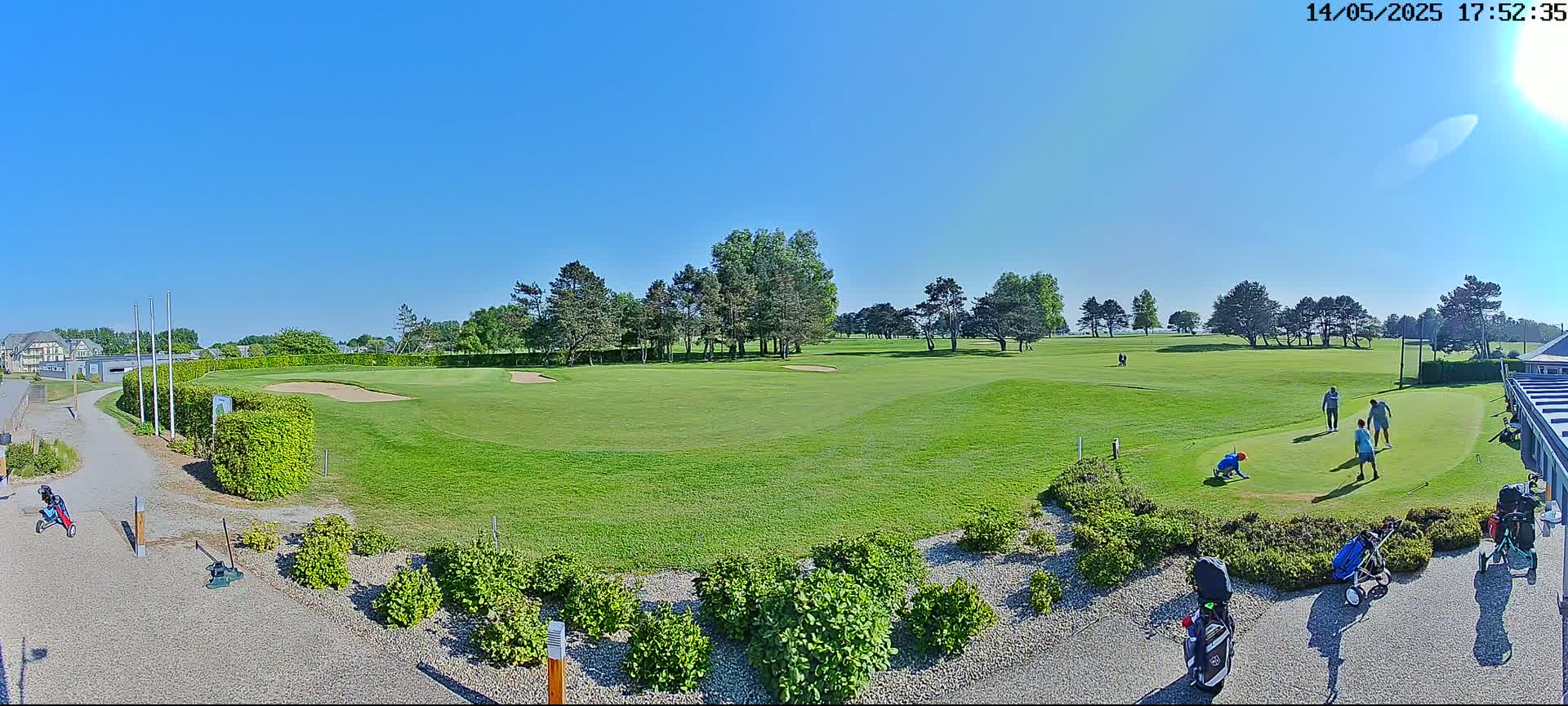 A sunny day at a golf course shows several golfers on the putting green, surrounded by green grass and trees.