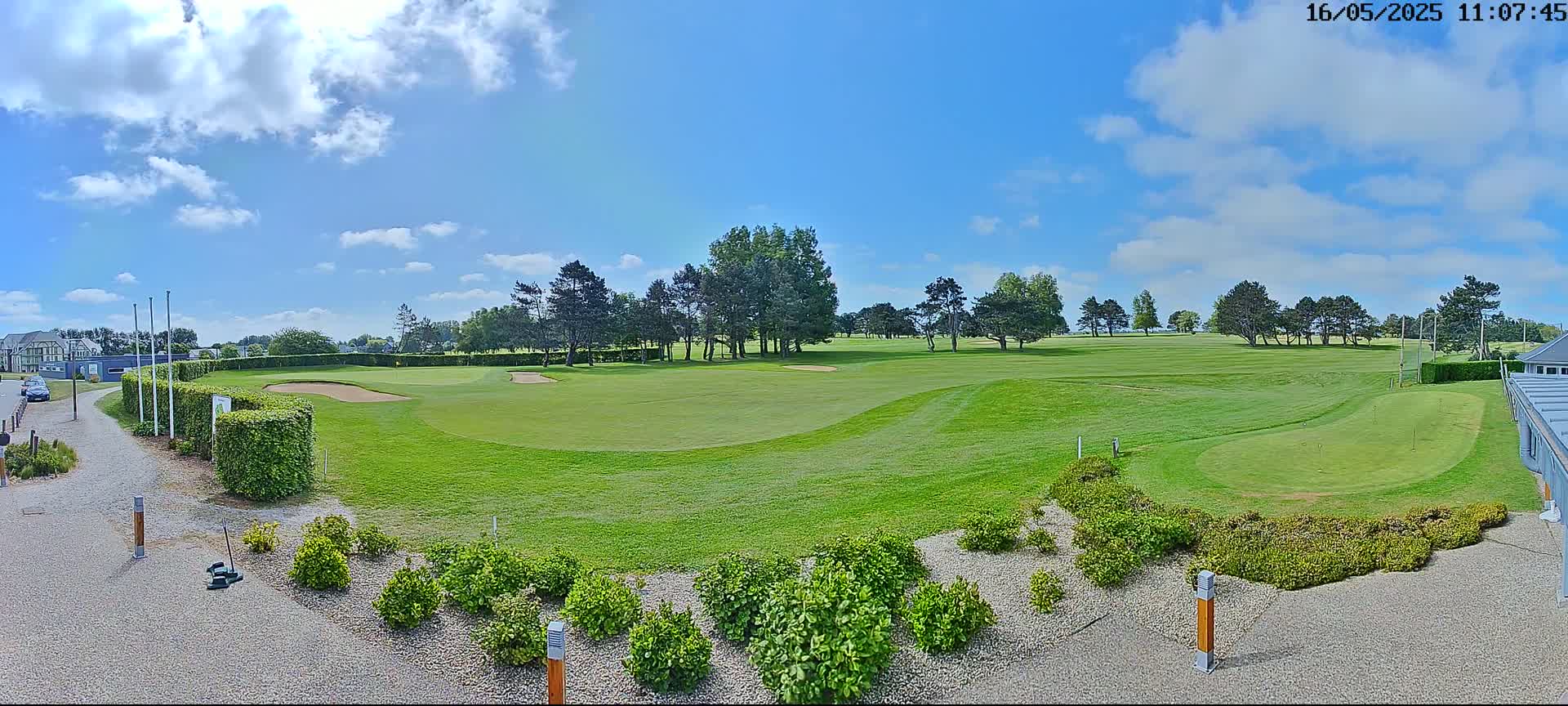 A sunny day on a golf course shows a putting green, sand traps, and neatly trimmed landscaping, with a mostly clear sky and a few fluffy clouds.