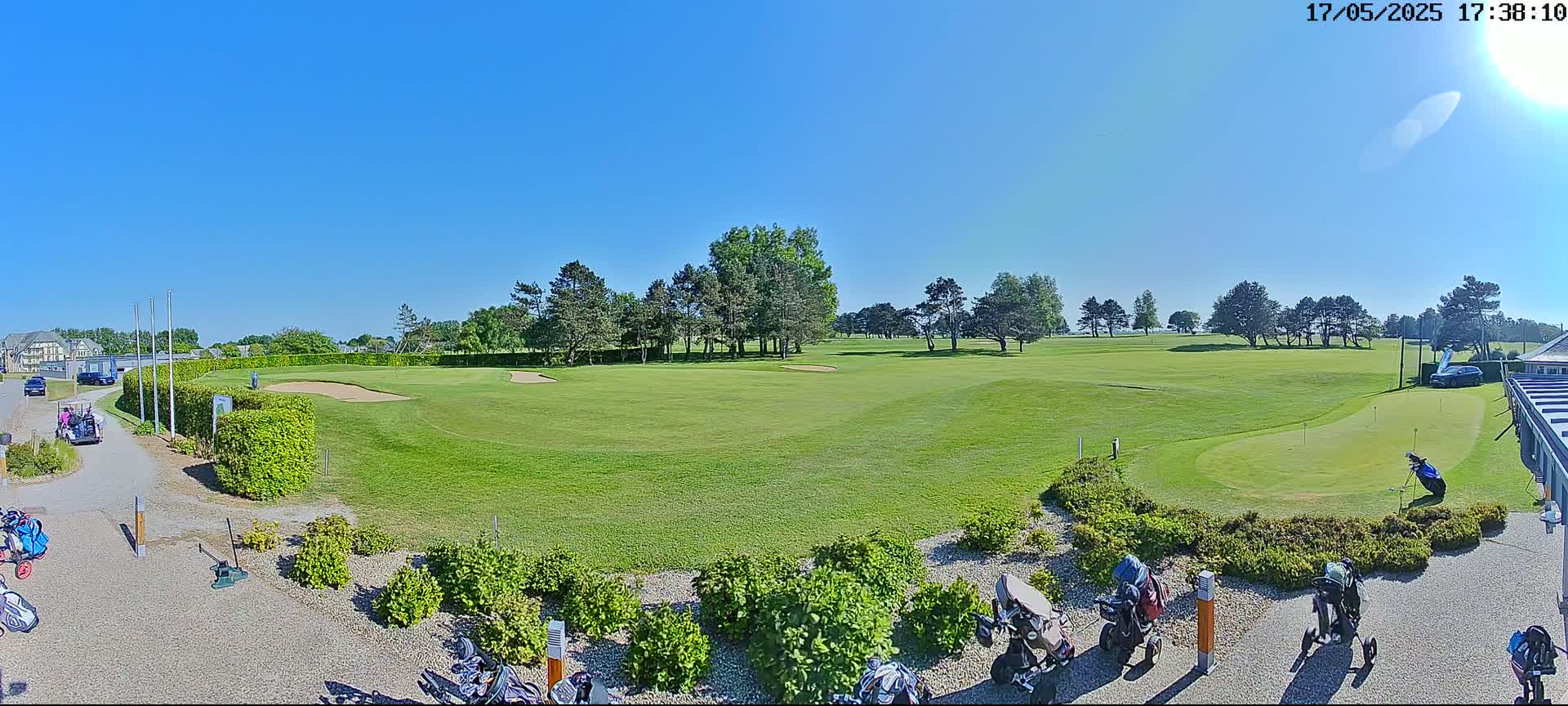 A sunny day on a golf course shows a putting green, several golf carts, and golfers near a clubhouse.