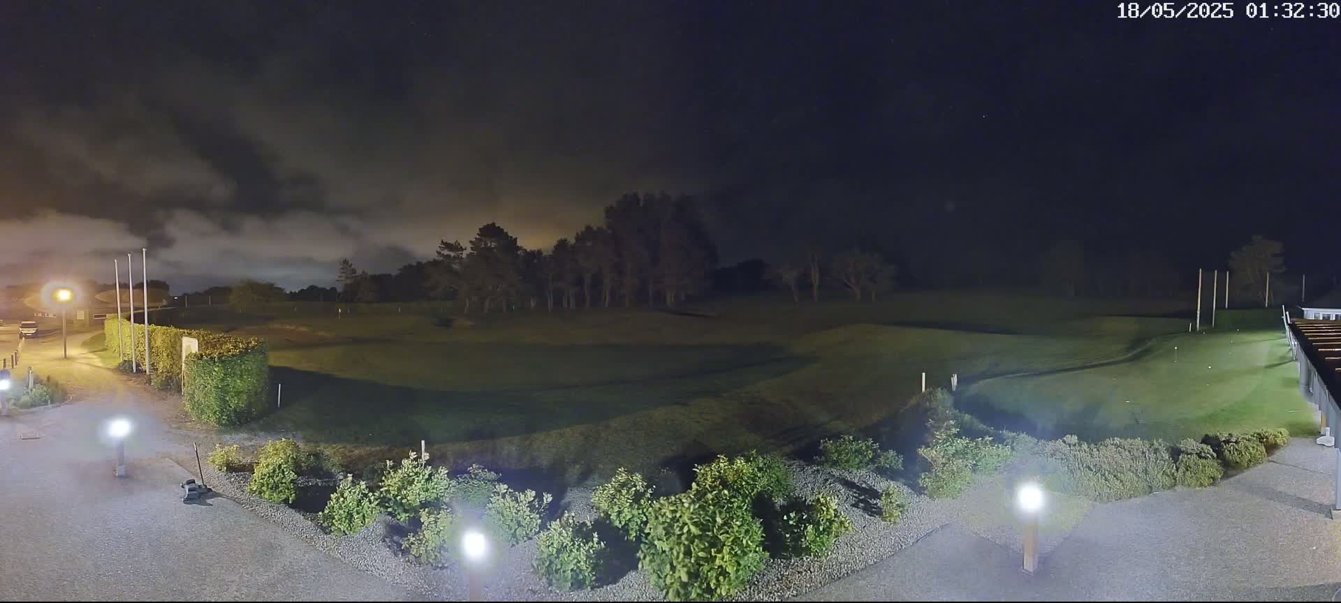 A nighttime view of a golf course under a cloudy sky, illuminated by several lights, shows a putting green and surrounding landscaping.