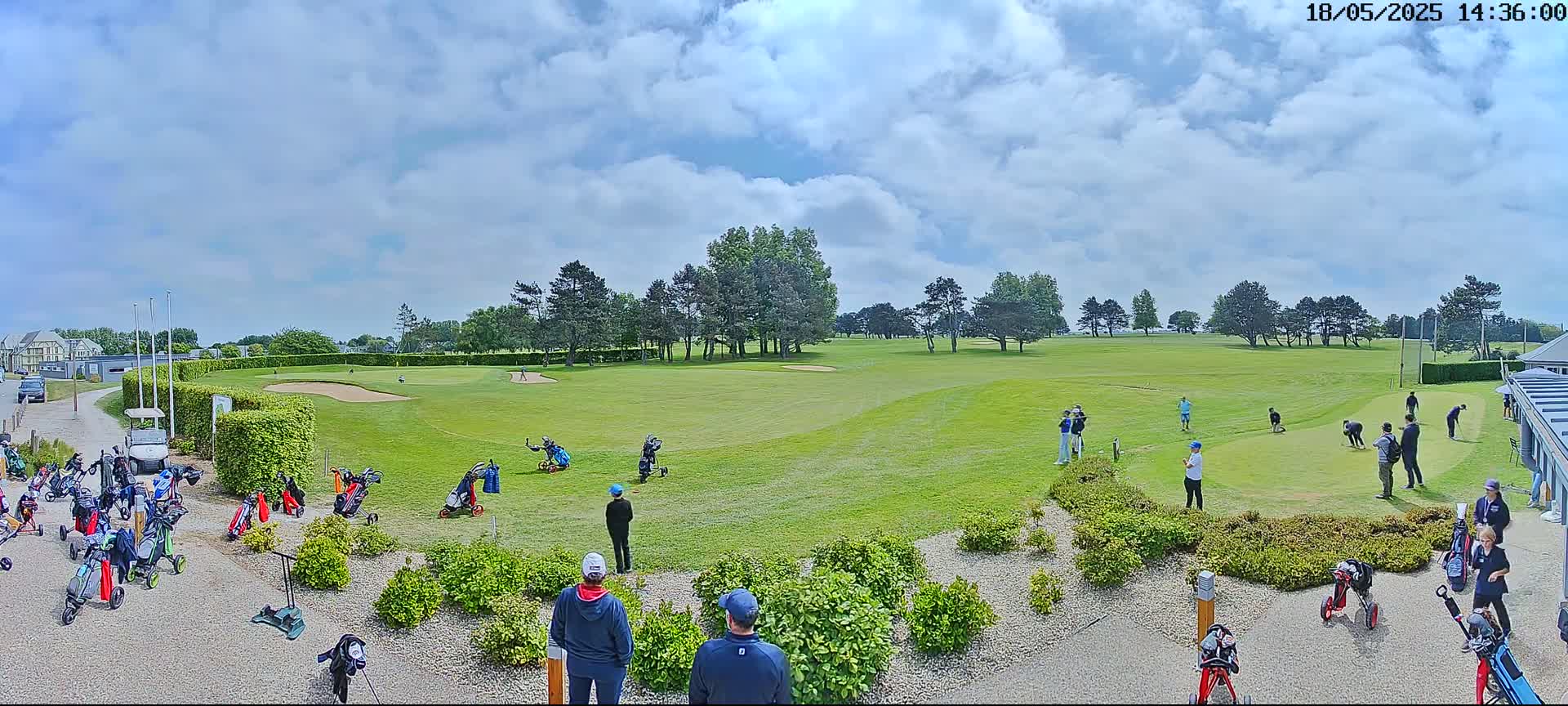 A sunny day at a golf course shows multiple golfers on the green and many golf carts parked near the clubhouse.