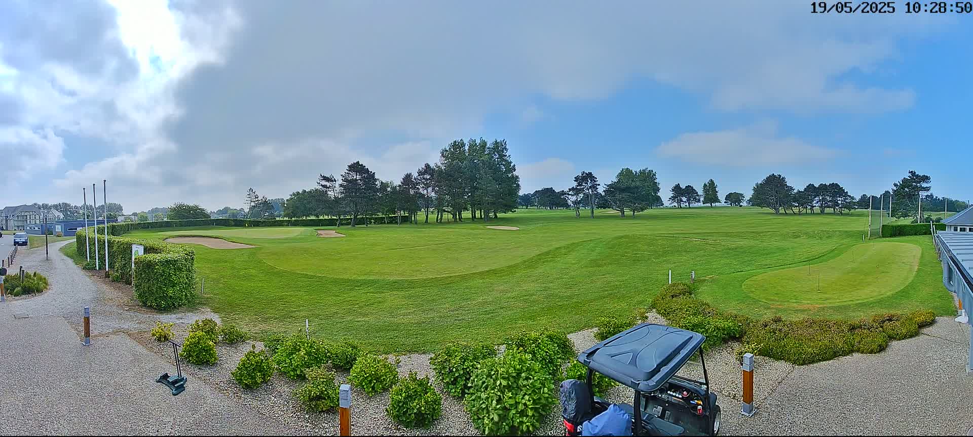 A partly cloudy sky overlooks a green golf course with a putting green, a golf cart, and landscaping.