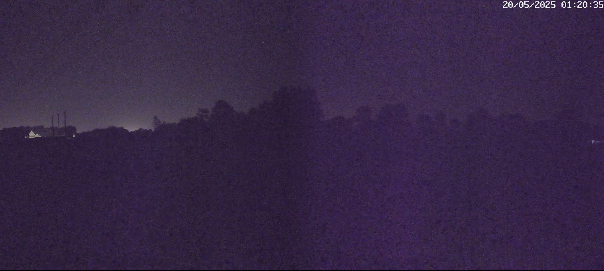 A nighttime view of a dark, flat landscape with a treeline and a few dimly lit structures in the distance under a dark sky.