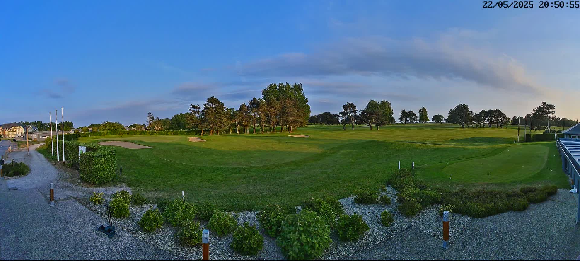 A panoramic view of a golf course under a mostly clear, blue sky shows a green fairway, sand traps, and landscaping near a building.