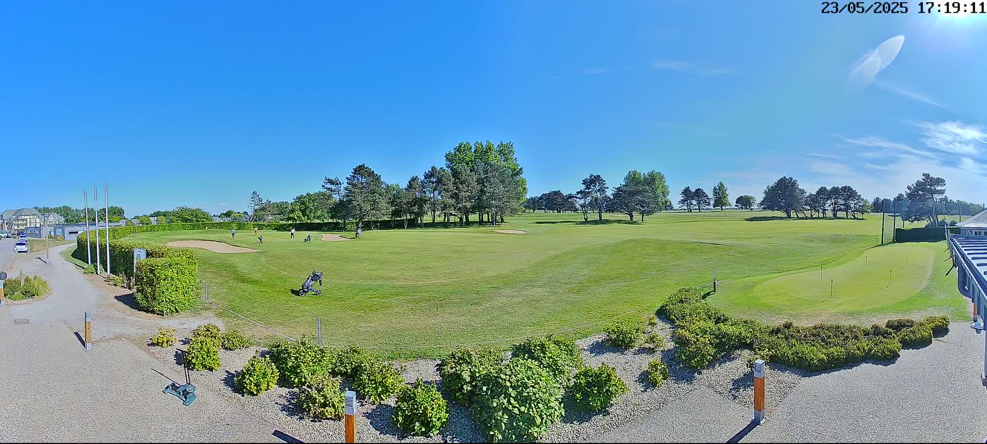 A sunny day on a golf course shows several golfers on a green, with a sand trap and putting green visible.
