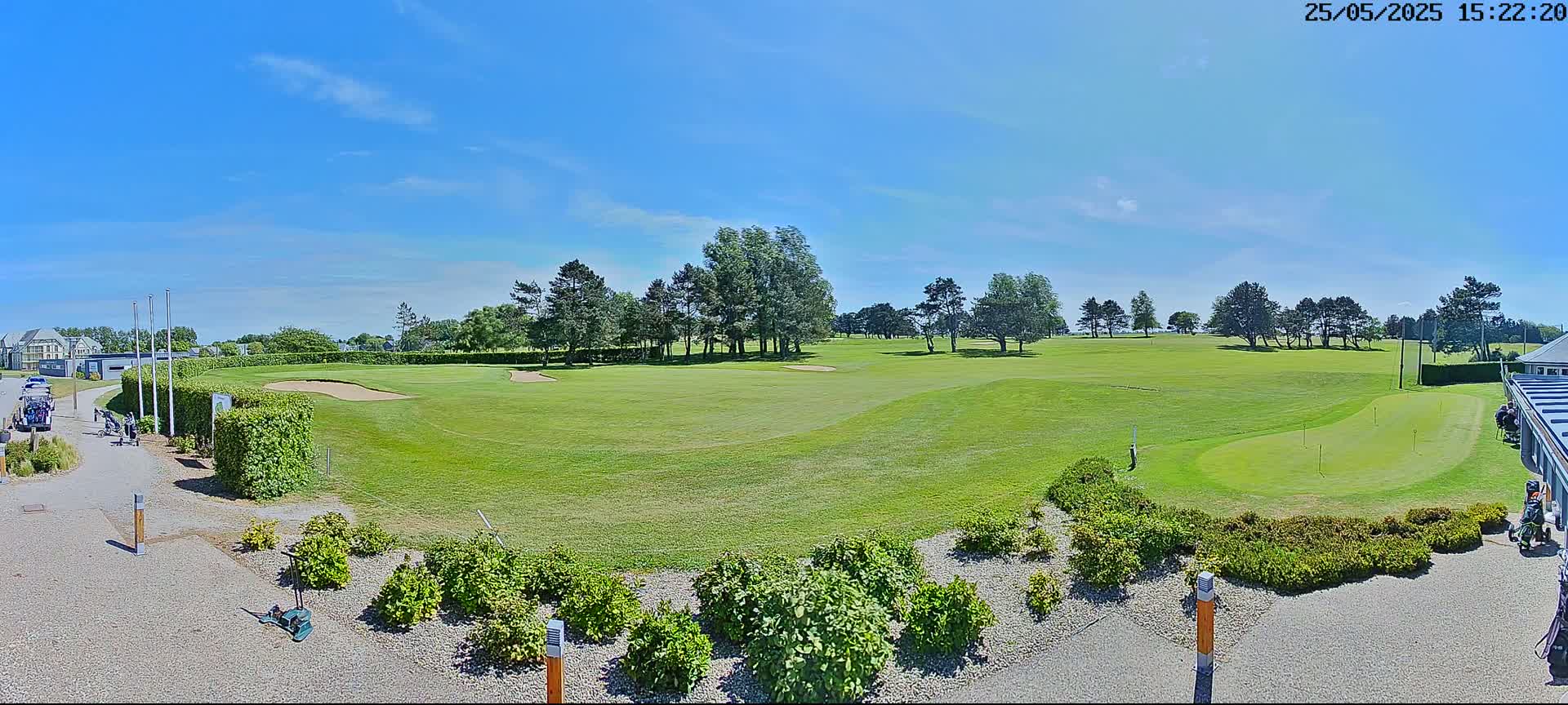 A sunny day on a golf course shows a green fairway, sand traps, and putting green, with trees lining the course and landscaping near the clubhouse.