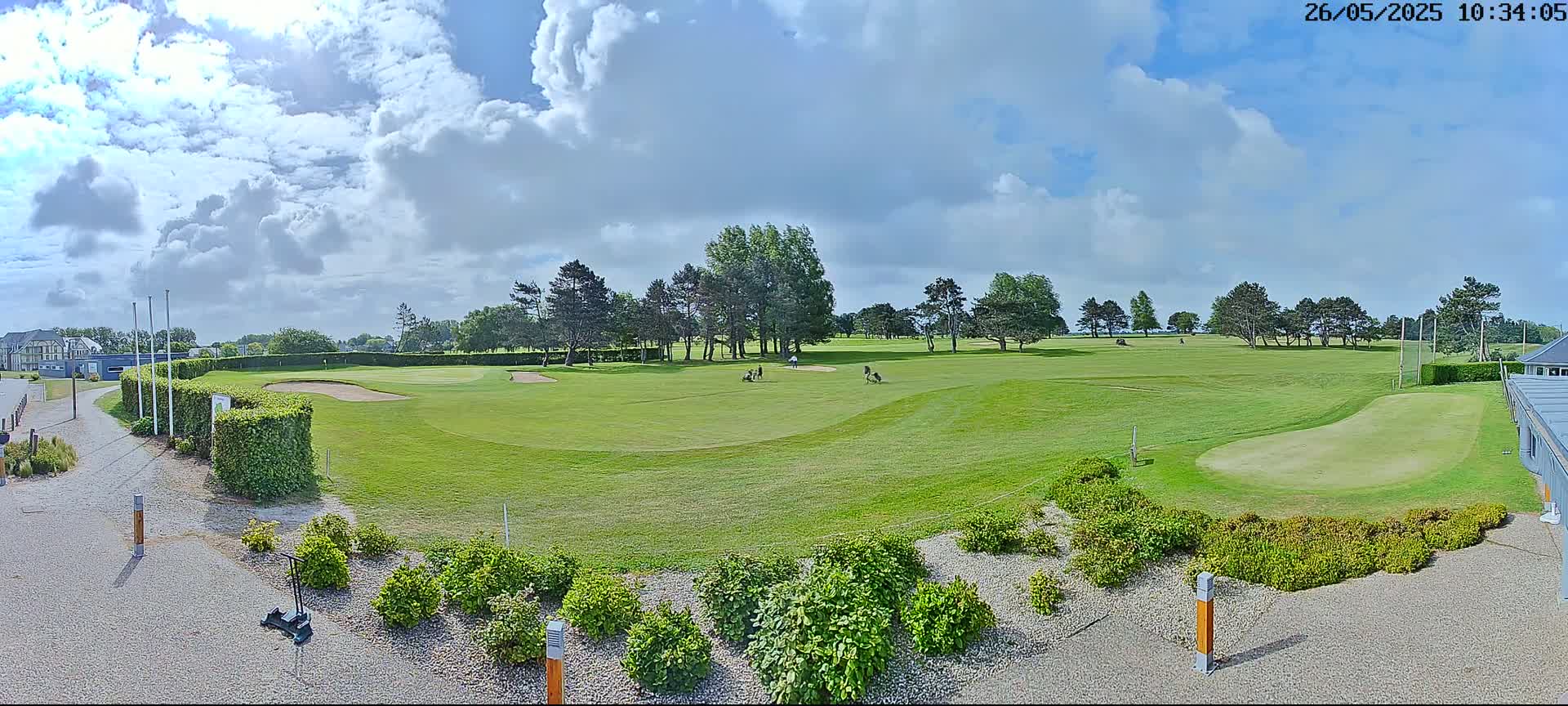 A mostly sunny day shows a golf course with a few golfers visible on the green, surrounded by trees and landscaping.