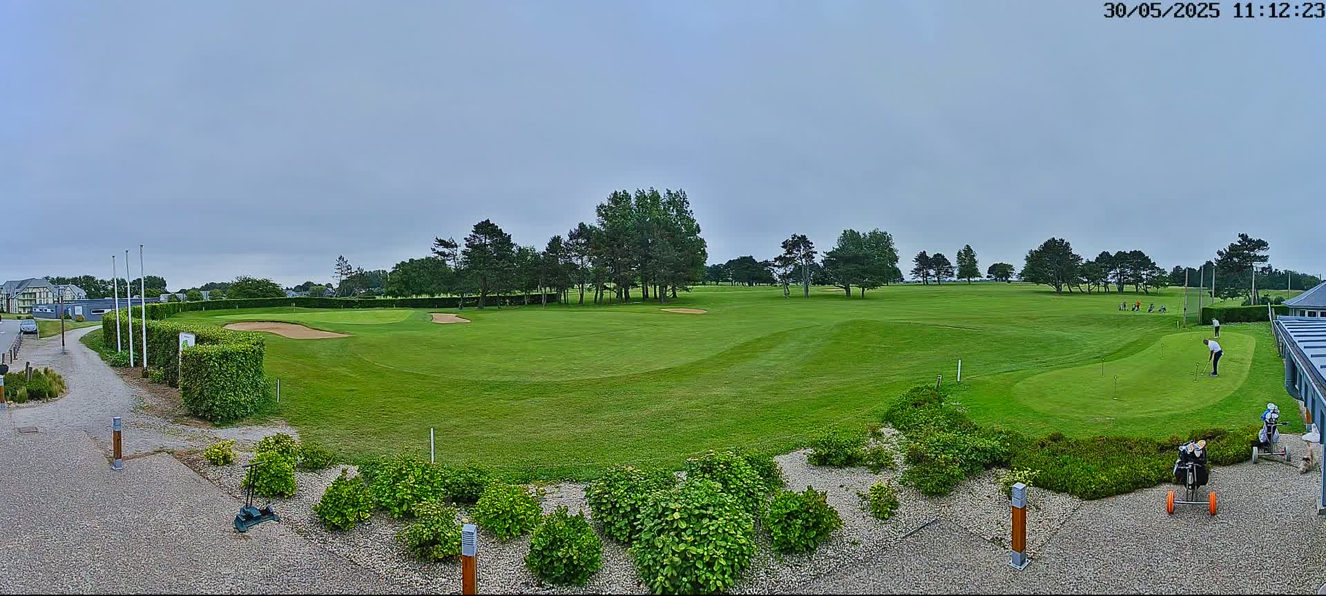 A green golf course with several golfers and golf carts is visible under an overcast sky.