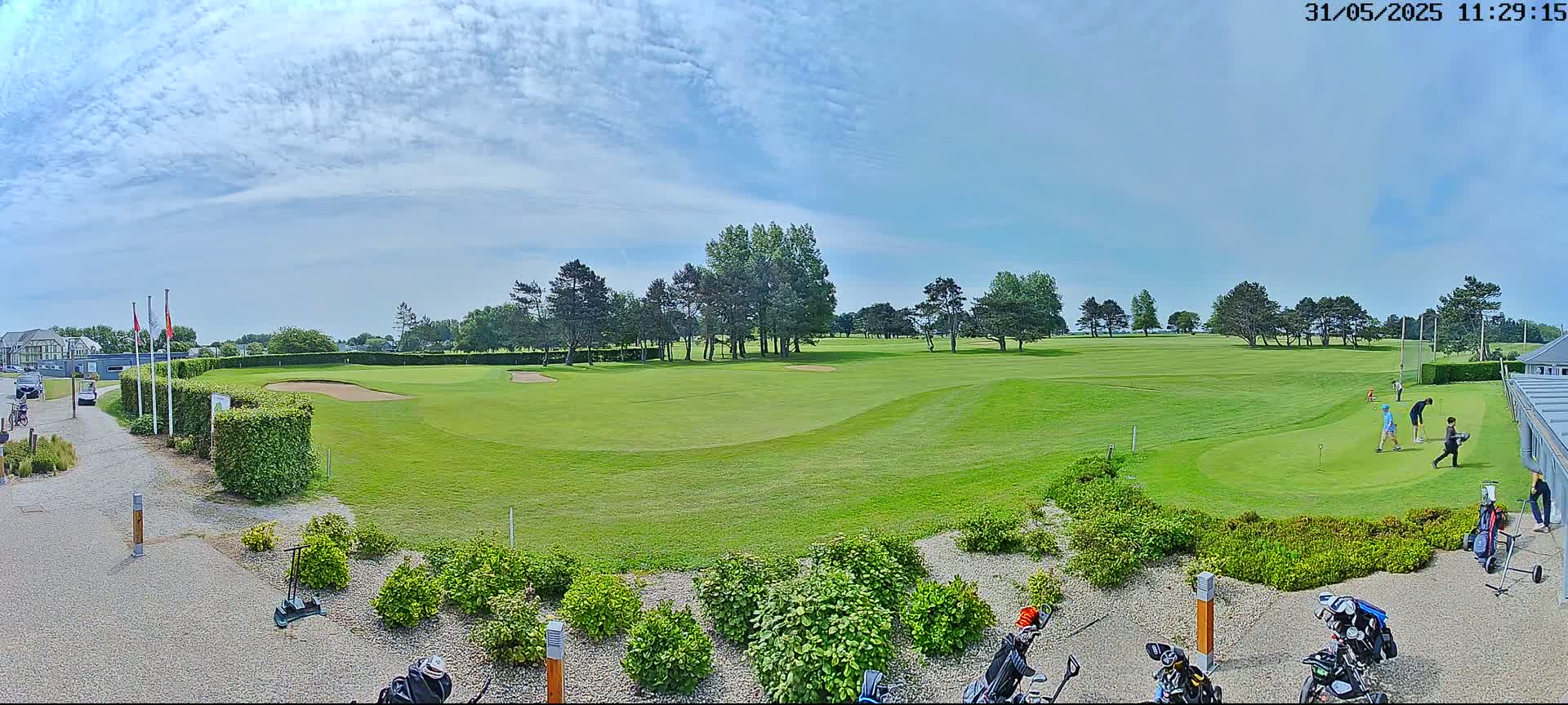 A sunny day on a golf course shows several golfers on the putting green, with golf bags and carts near a gravel pathway.