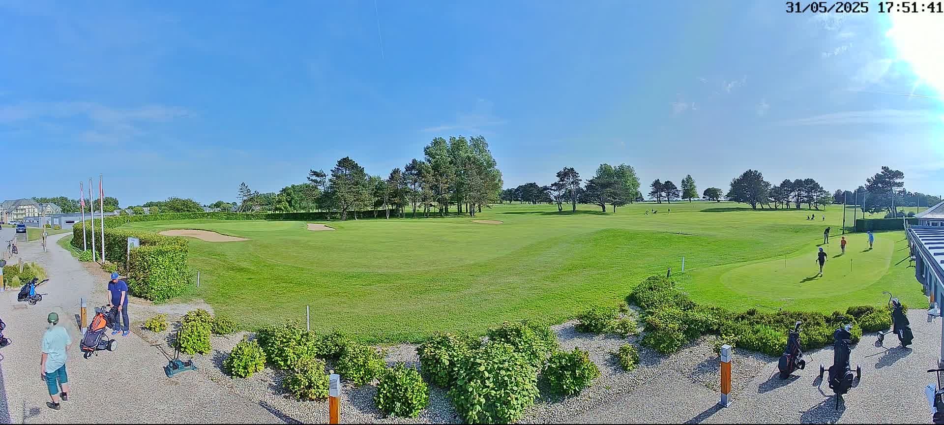 A sunny day on a golf course shows several people golfing on a green with trees and shrubs lining the course.