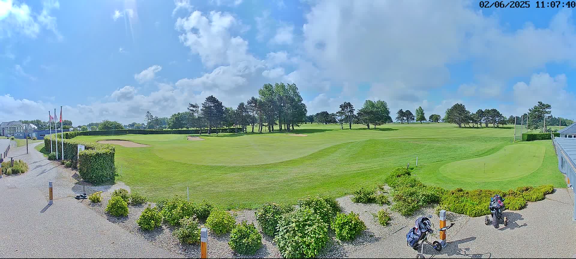 A sunny day shows a golf course with green fairways, sand traps, and a putting green, bordered by shrubs and trees.