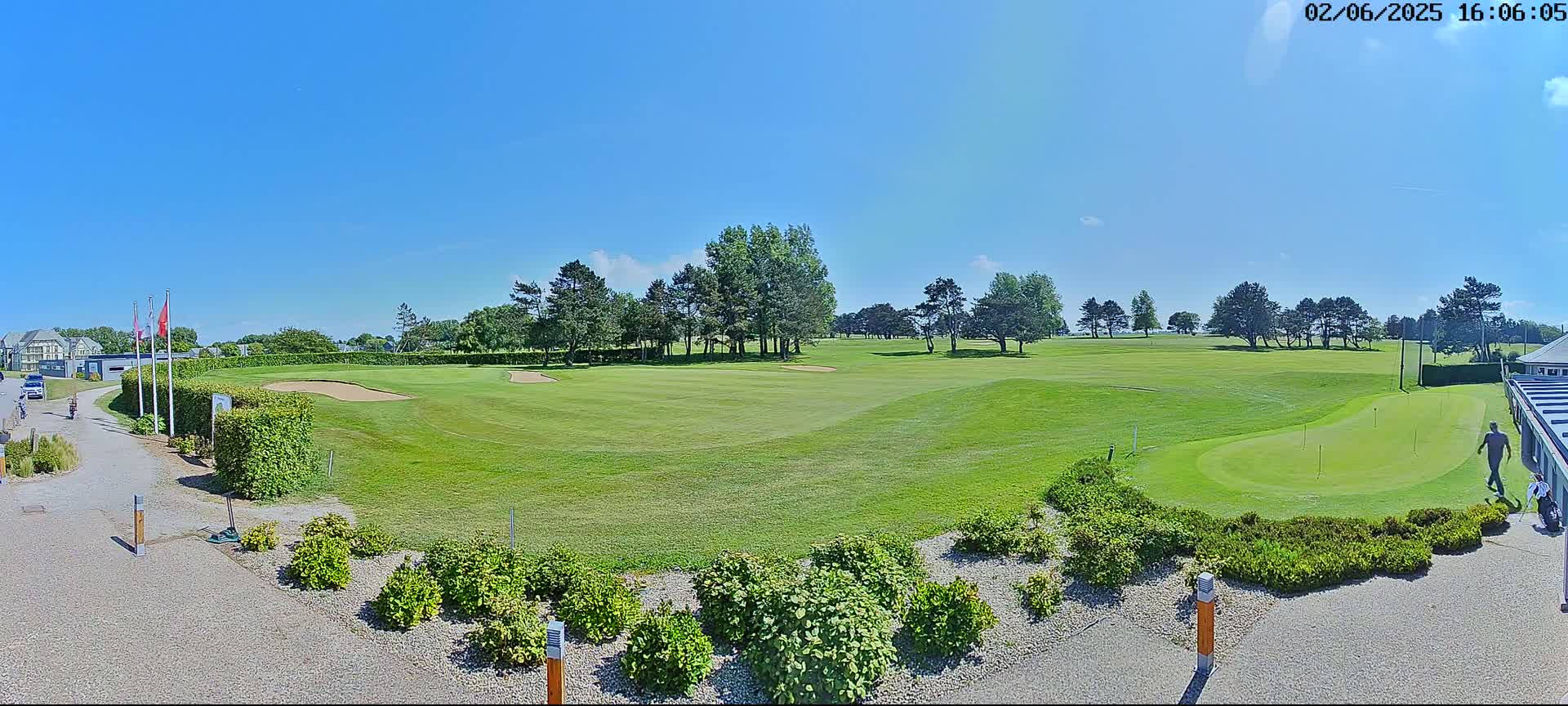 A sunny day on a golf course shows a putting green, a sand trap, and several people walking near a building.