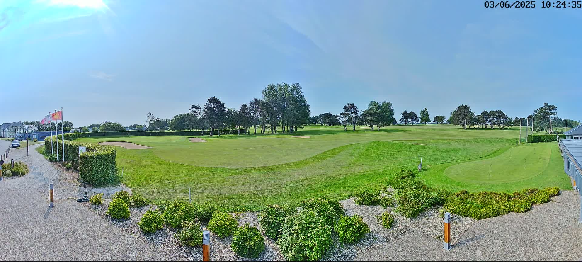 A sunny day on a golf course shows a green fairway, sand traps, and putting green, surrounded by trees and landscaping.