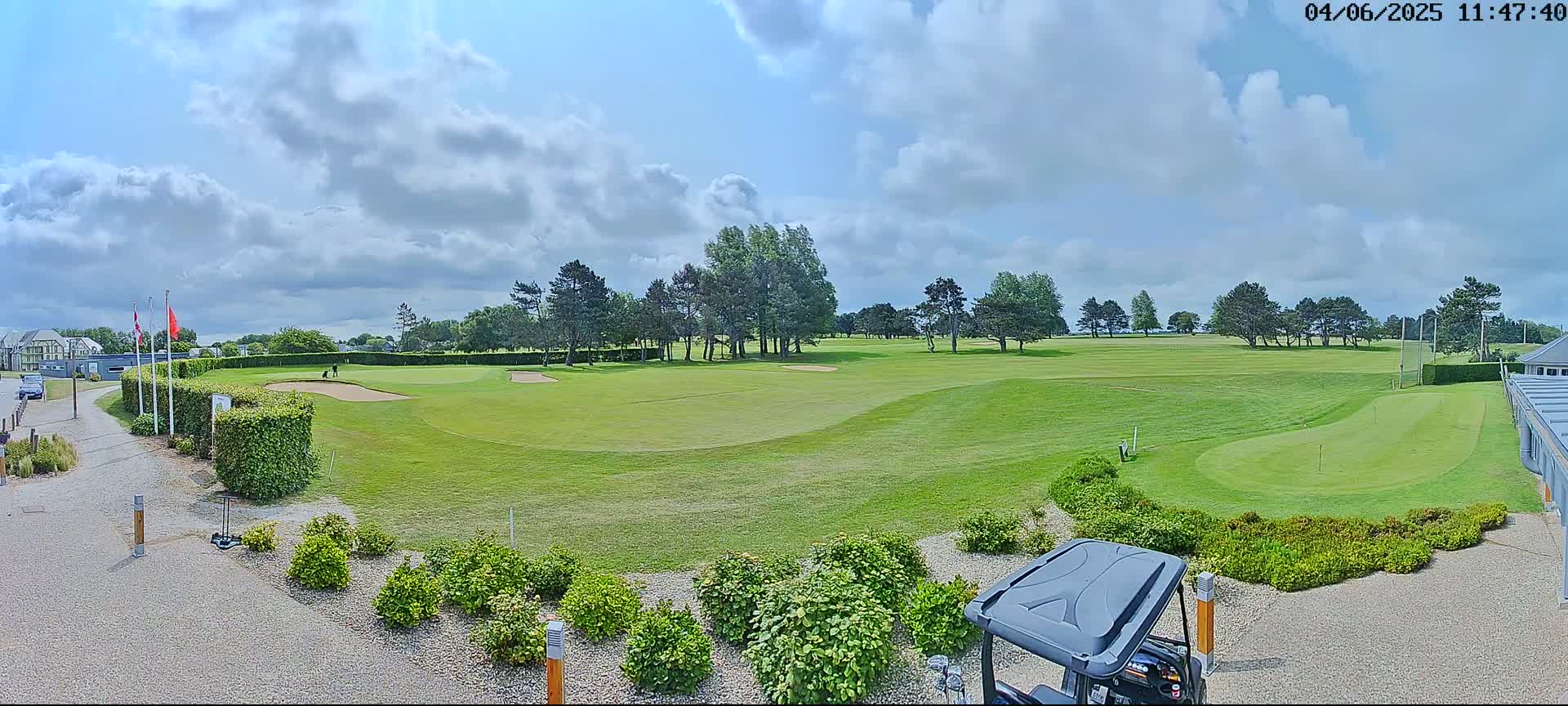 A partly cloudy day on a green golf course with a golf cart parked near lush bushes.