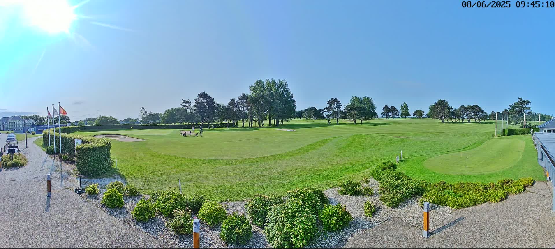 A sunny day on a golf course shows a putting green in the foreground, a fairway extending into the distance, and several golfers playing on the course.