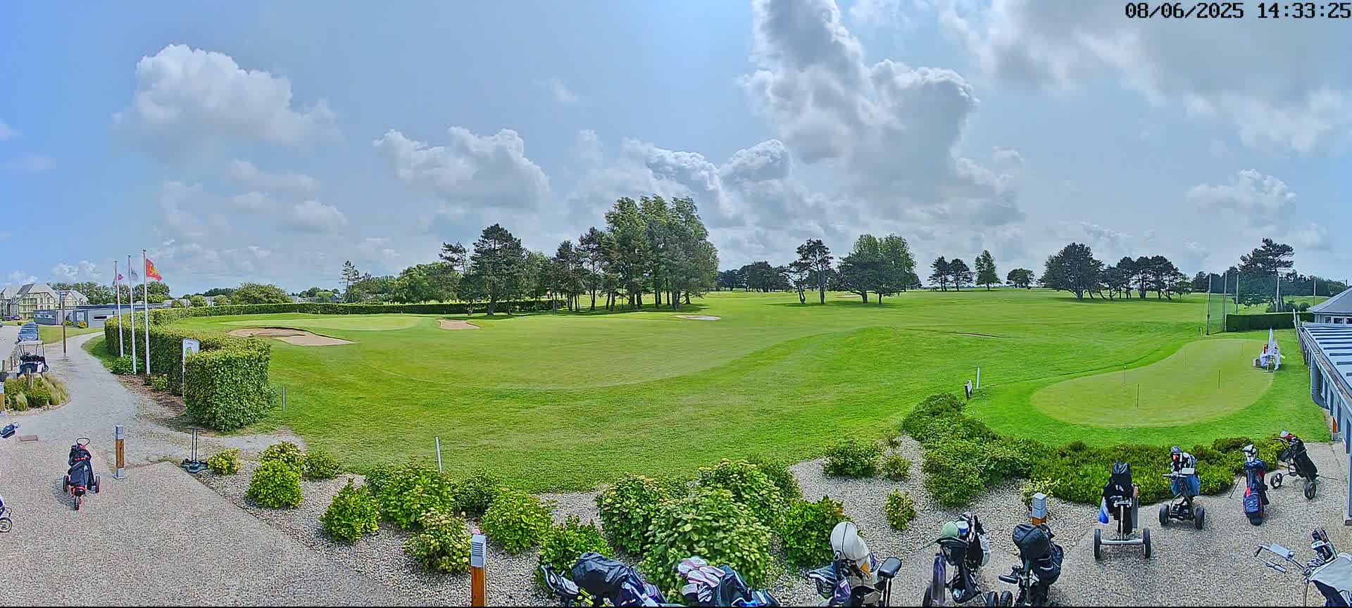 A sunny day on a golf course shows a putting green, several golf carts, and bags of golf clubs near a building.