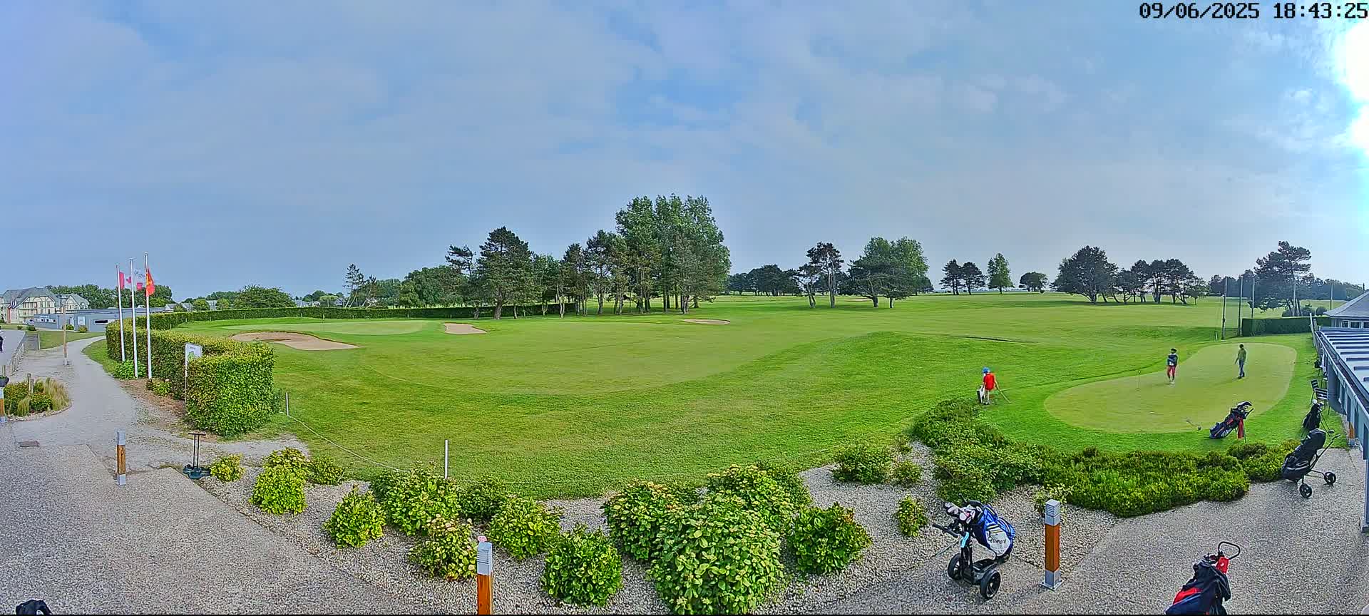 A mostly sunny day on a golf course shows several golfers on a green, with additional golfers and golf carts visible nearby.