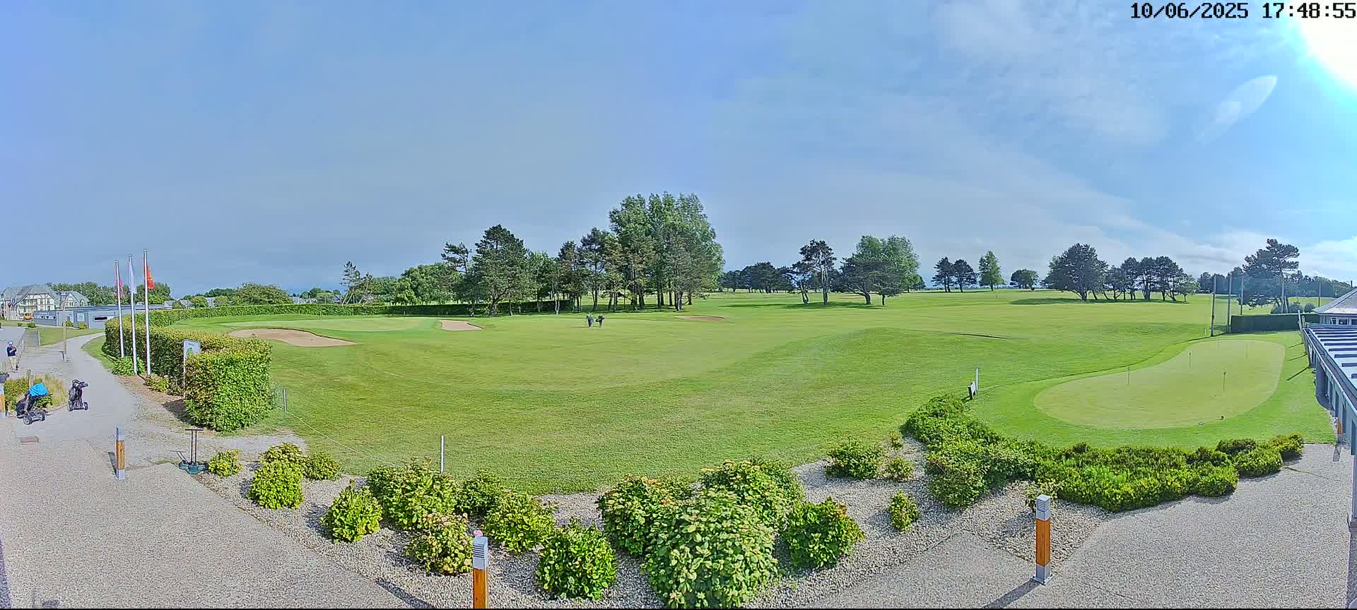 A sunny day reveals a golf course with a putting green in the foreground, several golfers visible in the distance, and landscaping surrounding the paved walkway.