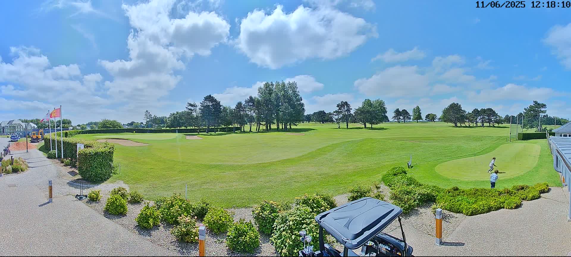 A sunny day on a golf course shows a putting green with two people, a golf cart, and a partly cloudy blue sky.