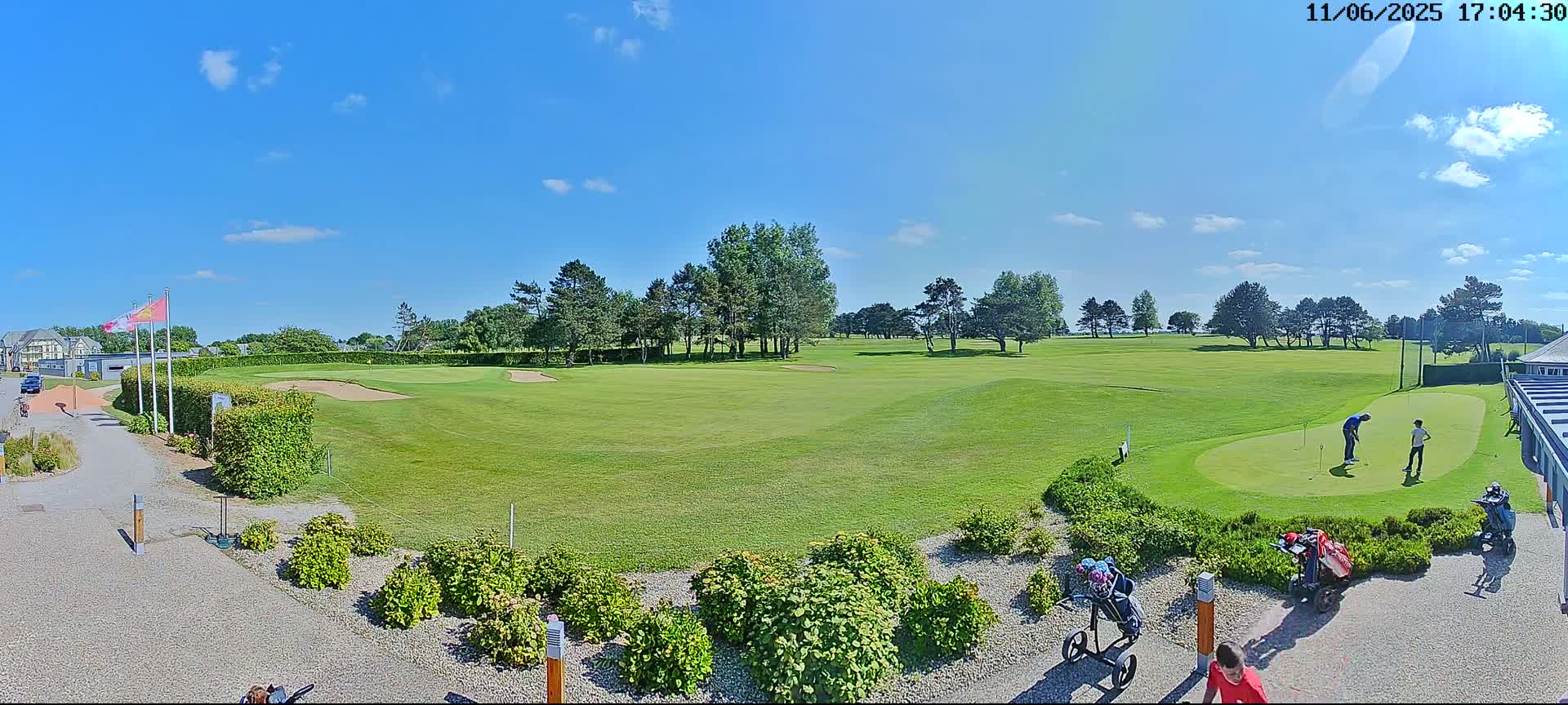 A sunny day on a golf course shows a green putting green with golfers, bordered by landscaping and trees under a clear blue sky.