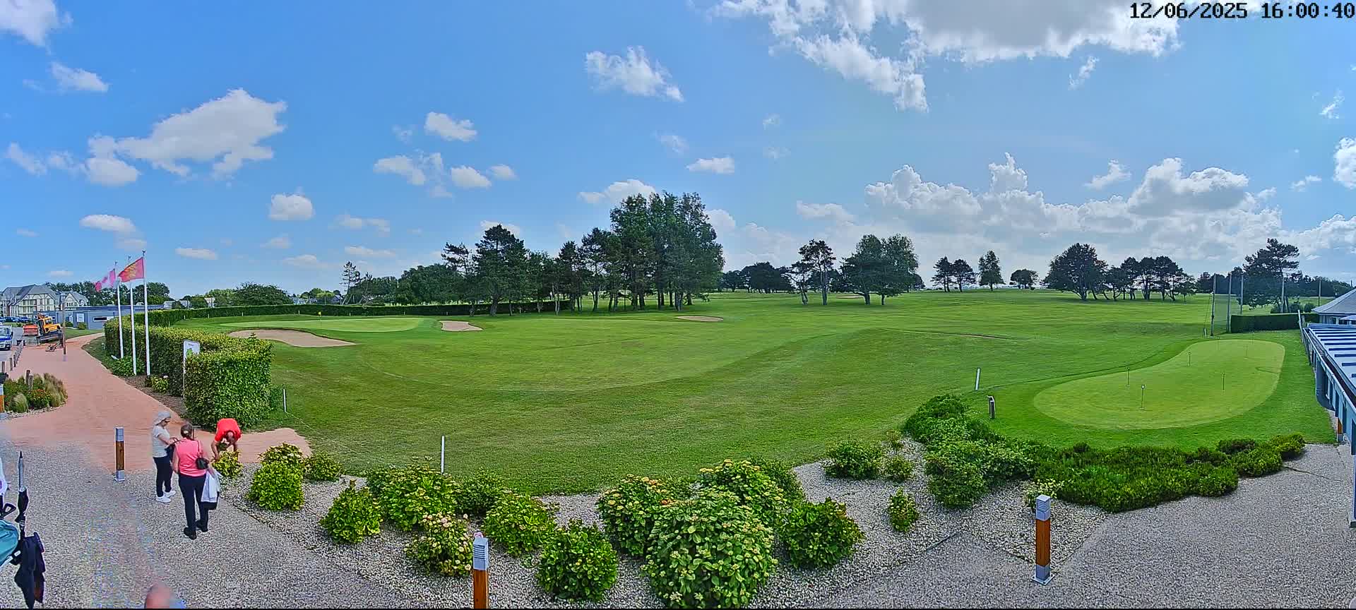 A sunny day with some clouds shows a green golf course with people near the clubhouse and a putting green in the foreground.